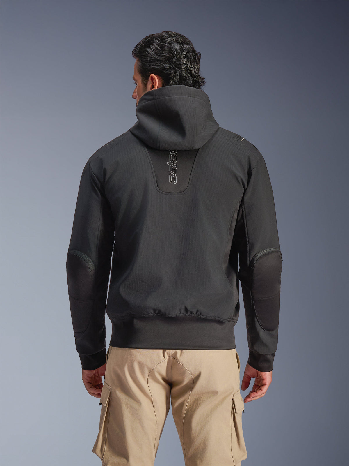 Alpinestars Moblast Waterproof Jacket, czarna, miejska kurtka motocyklowa z kapturem, widok z tyłu, pionowe logo „astars” na plecach, elastyczny pas i mankiety, wzmocnione łokcie, nowoczesny design, beżowe spodnie cargo