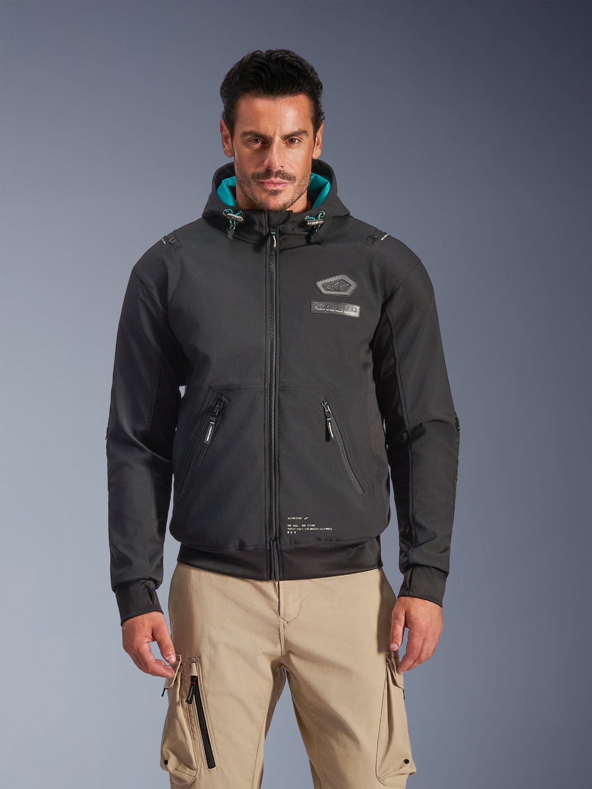 Alpinestars Moblast Waterproof Jacket, męska miejska kurtka motocyklowa, czarna z turkusową podszewką kaptura, posiada kieszenie 3D na zamek i fason z kapturem, noszona z beżowymi spodniami cargo, nowoczesna miejska odzież motocyklowa