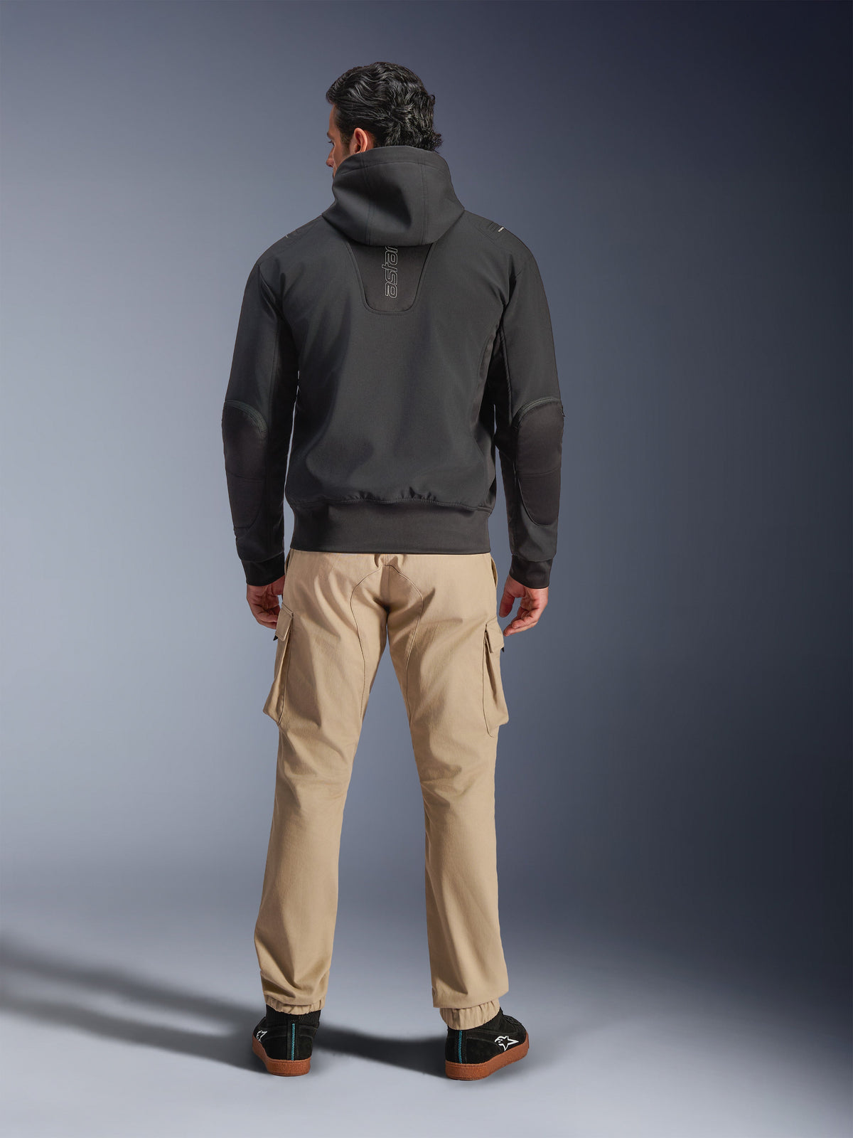 Wodoodporna kurtka Alpinestars Moblast, czarna, widok z tyłu miejskiej bluzy motocyklowej, zestawiona z beżowymi spodniami cargo i czarnymi sneakersami, minimalistyczny design z pionowym napisem „astars” w górnej części pleców, nowoczesna odzież motocyklowa na każdą pogodę