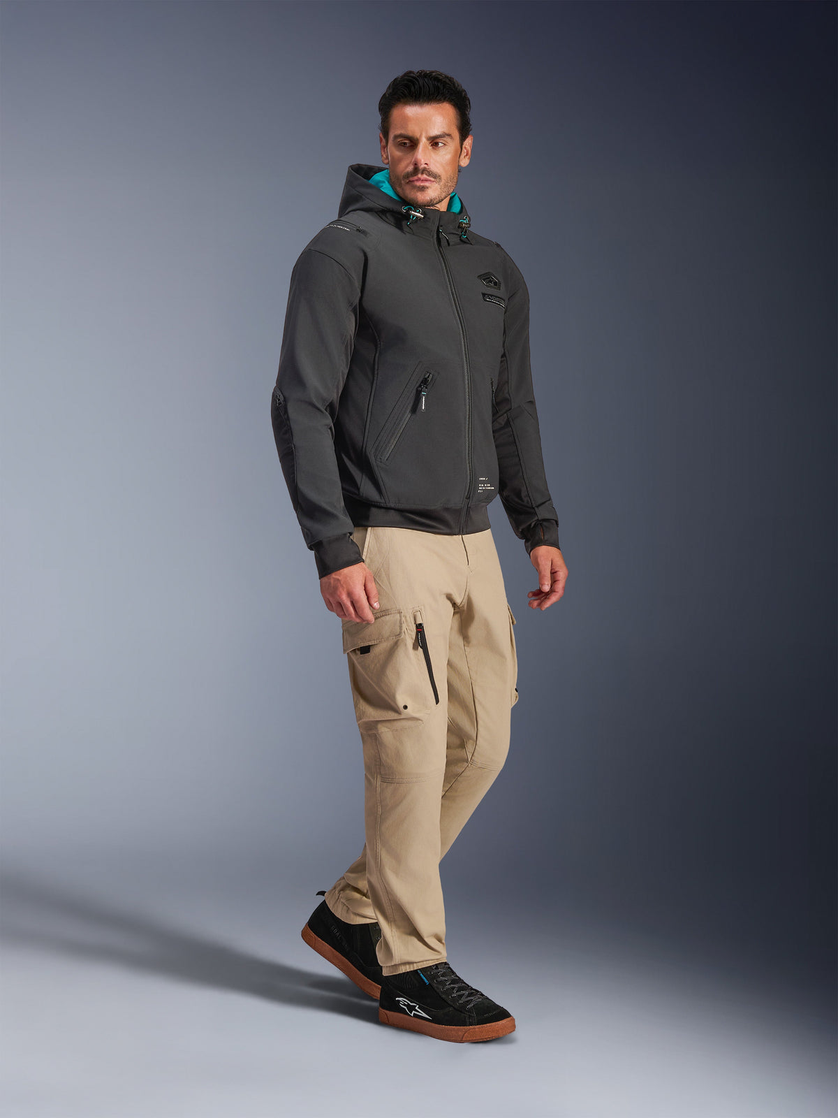 Męski model ubrany w czarną kurtkę Alpinestars Moblast Waterproof Jacket, miejska bluza motocyklowa z kapturem z turkusową podszewką, w zestawieniu ze spodniami cargo khaki i czarnymi sneakersami, nowoczesny minimalistyczny design z technicznego stretchu na każdą pogodę