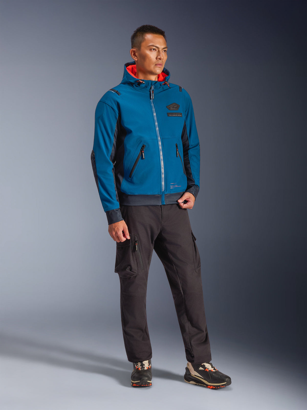 Male model wearing the Alpinestars Moblast Waterproof Jacket in Dark Petrol and Dark Navy, featuring a hooded design with a bright orange interior, paired with black cargo pants and sneakers, modern urban motorcycle riding gear.