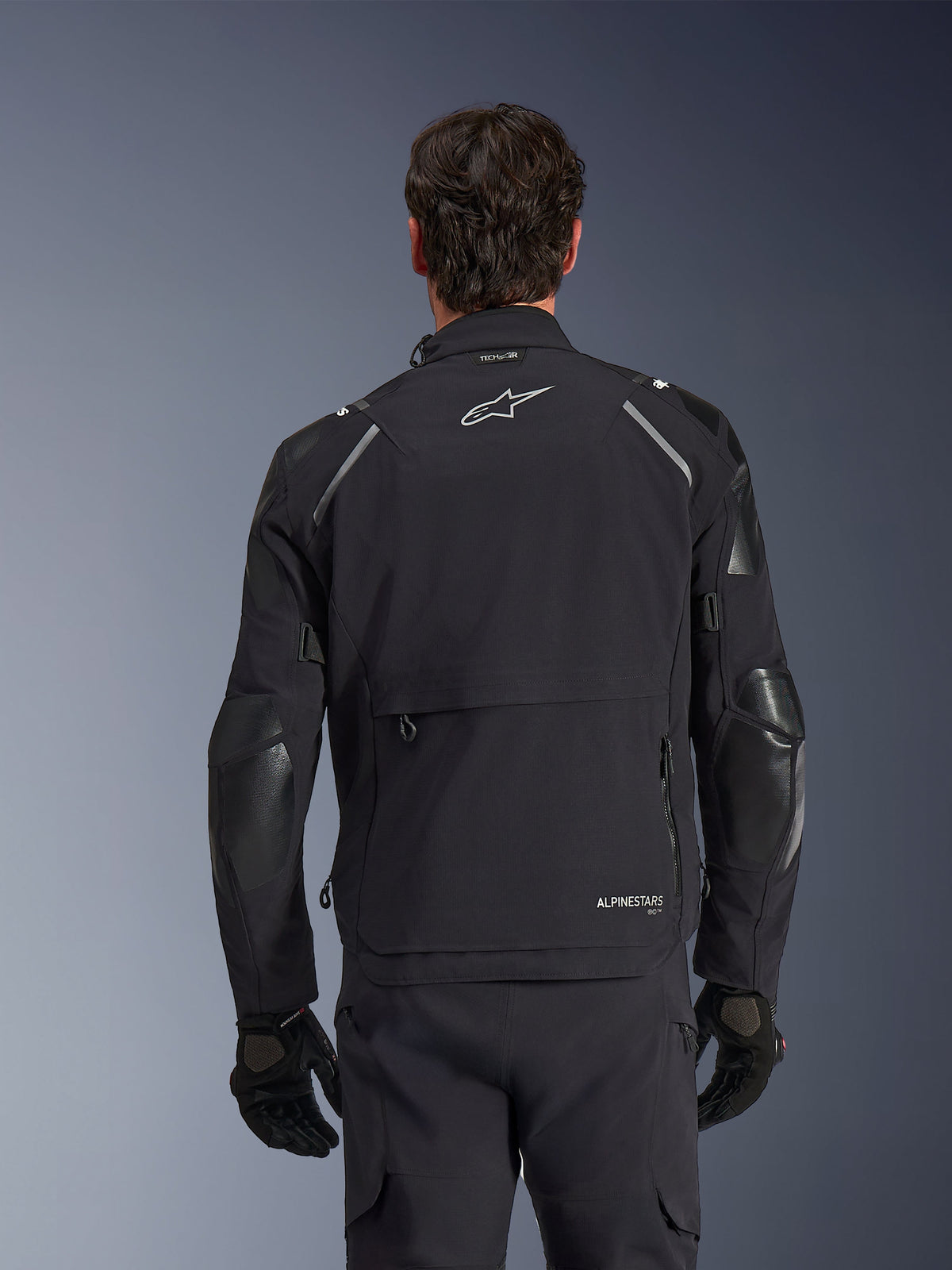 Kurtka Alpinestars Andes Pro Drystar® XF, czarno-ciemnoszara, widok z tyłu na modelu, wyposażona w dużą tylną komorę do przechowywania, odblaskowe detale na ramionach i logo, zestawiona z czarnymi rękawicami i spodniami do jazdy typu adventure.