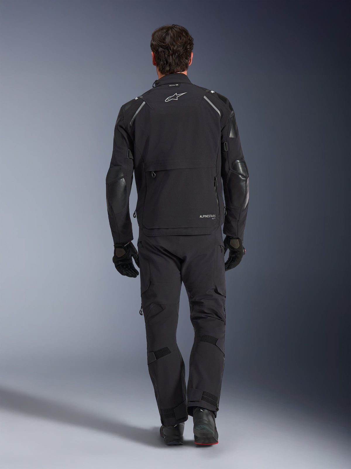 Kurtka Alpinestars Andes Pro Drystar® XF, kurtka adventure touring, czarna i ciemnoszara, widok z tyłu pokazujący dużą kieszeń cargo i wzmocnione panele na łokciach, noszona z dopasowanymi czarnymi spodniami i rękawicami, funkcjonalny design do jazdy w każdych warunkach pogodowych