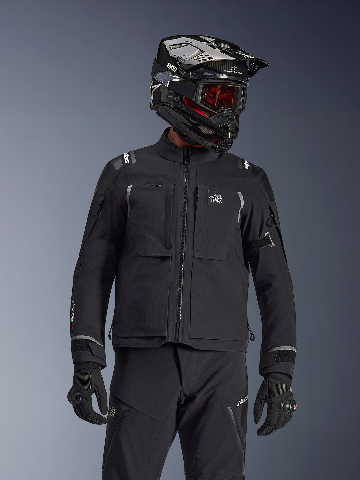Motocyklista adventure w kurtce Alpinestars Andes Pro Drystar® XF w kolorze czarnym i ciemnoszarym, z logo All Terra na klatce piersiowej, w zestawie z dopasowanymi spodniami technicznymi i czarnym kaskiem adventure z goglami, profesjonalny komplet odzieży turystycznej.