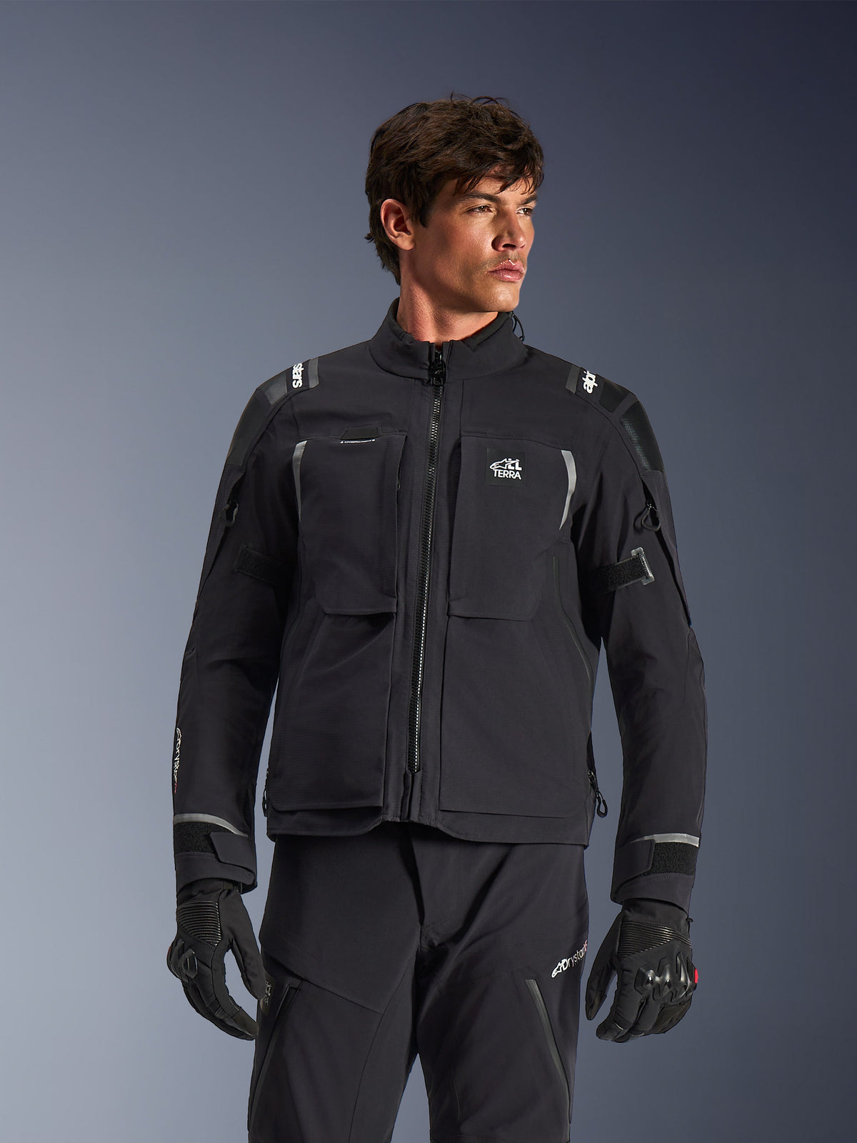 Alpinestars Andes Pro Drystar® XF Jacket, czarno-ciemnoszara motocyklowa kurtka turystyczna adventure, noszona z pasującymi spodniami i rękawicami, charakteryzująca się techniczną laminowaną konstrukcją zewnętrzną, logo ALL TERRA na klatce piersiowej, odblaskowymi detalami i regulowanymi mankietami