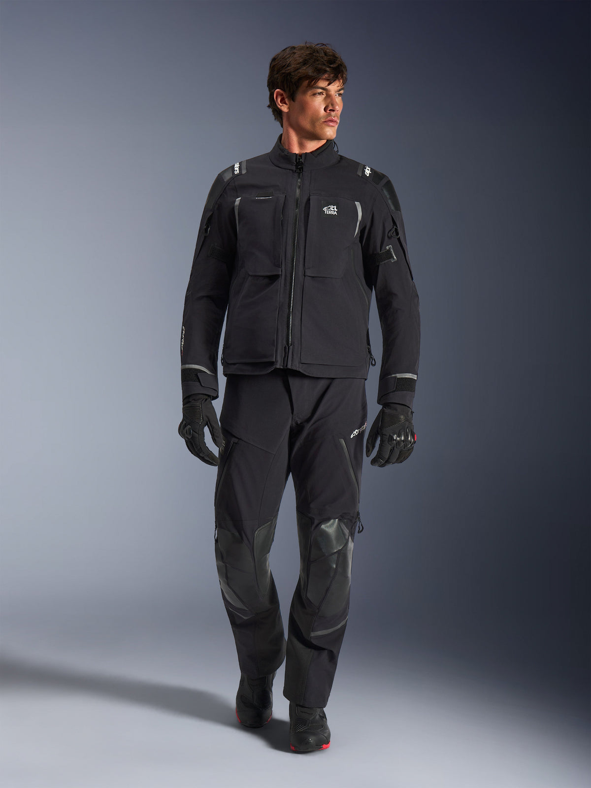 Męski model w kurtce Alpinestars Andes Pro Drystar® XF, czarno-ciemnoszara motocyklowa kurtka adventure, w zestawie z pasującymi spodniami technicznymi i rękawicami, z wieloma kieszeniami i brandingiem „Terra” na klatce piersiowej.