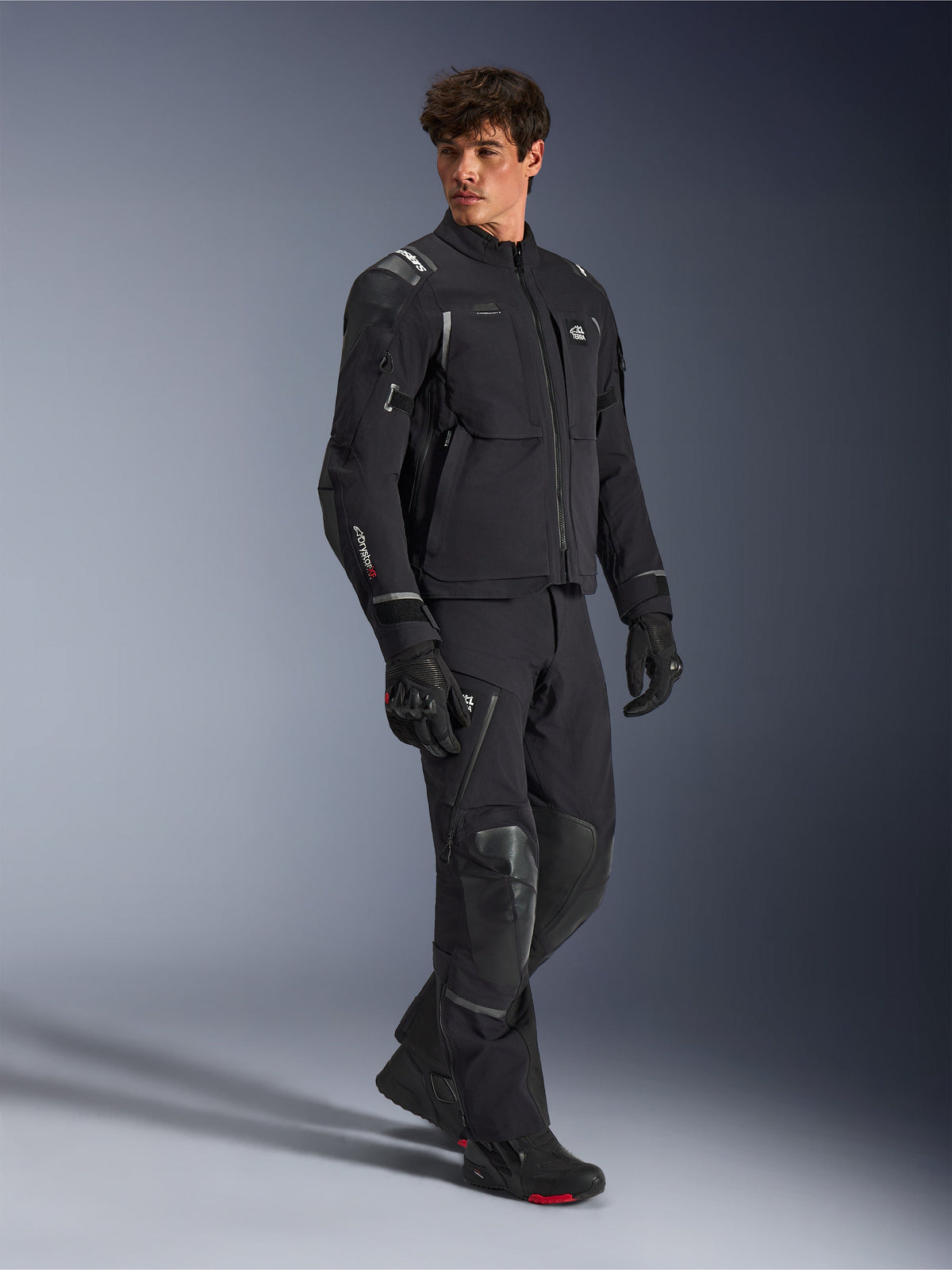 Kurtka Alpinestars Andes Pro Drystar® XF, czarno-ciemnoszara kurtka motocyklowa, noszona przez modela z dopasowanymi spodniami adventure, rękawicami i butami, ujęcie studyjne całej sylwetki, techniczna odzież turystyczna z odblaskowymi detalami i brandingiem