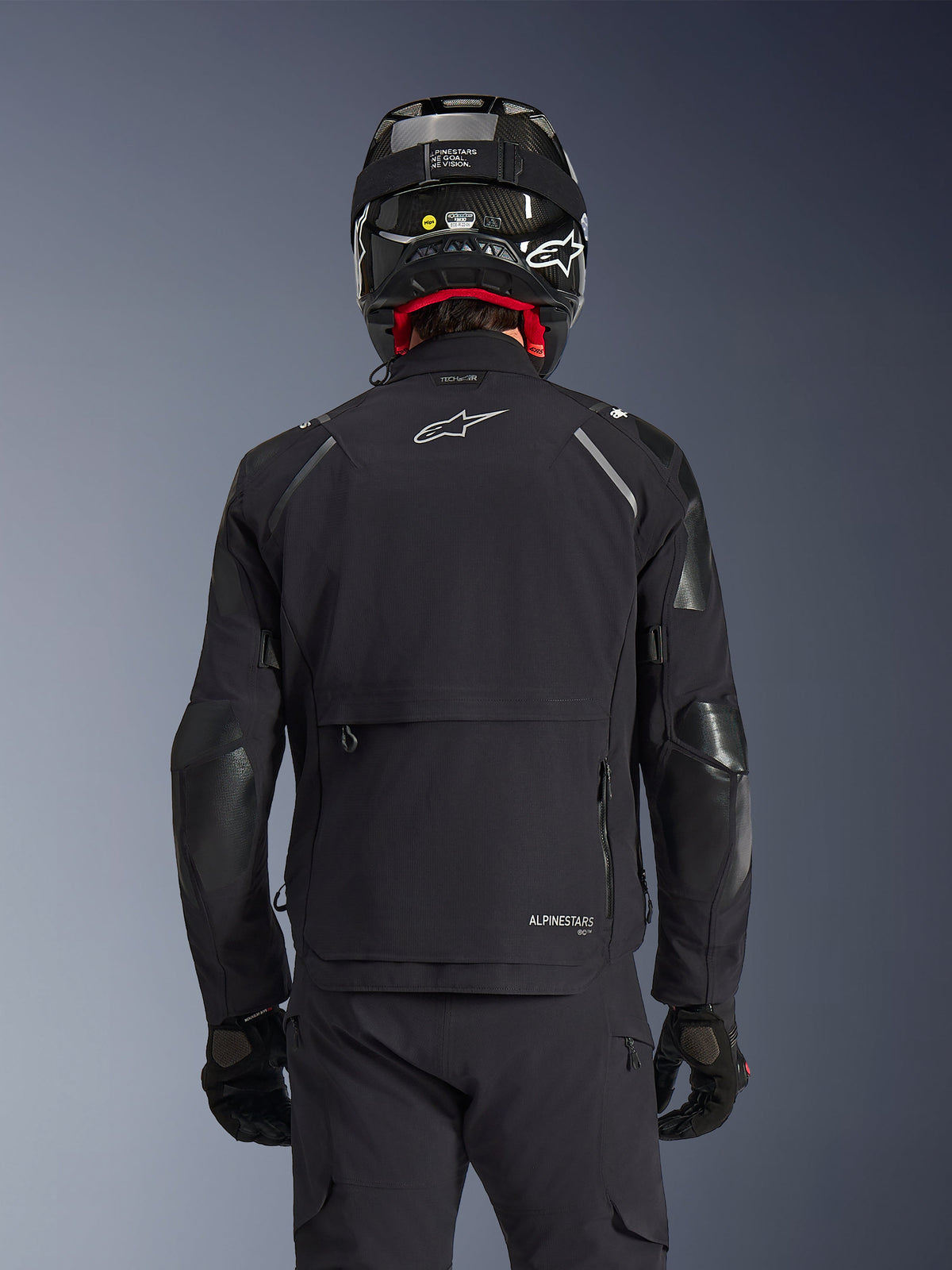 Kurtka Alpinestars Andes Pro Drystar® XF w kolorze czarnym, widok z tyłu na motocyklistę w kasku z włókna węglowego, duża tylna kieszeń i białe logo Alpinestars, techniczna całosezonowa odzież motocyklowa adventure