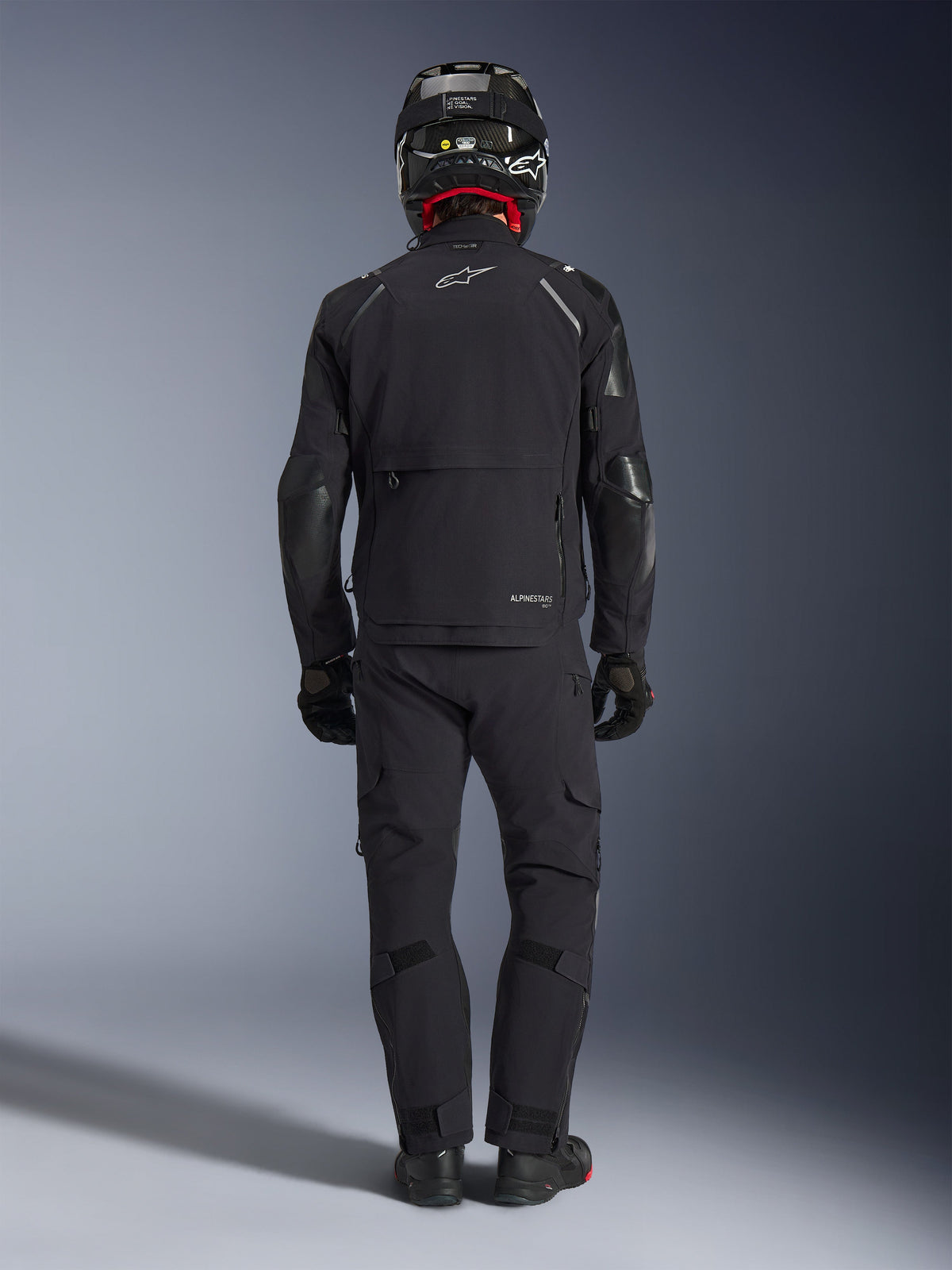 Kurtka Alpinestars Andes Pro Drystar® XF w kolorze czarnym i ciemnoszarym, widok z tyłu na motocyklistę w pełnym stroju adventure touring, pasujące spodnie i kask z włókna węglowego, duża tylna kieszeń cargo oraz odblaskowe logo Alpinestars na plecach.