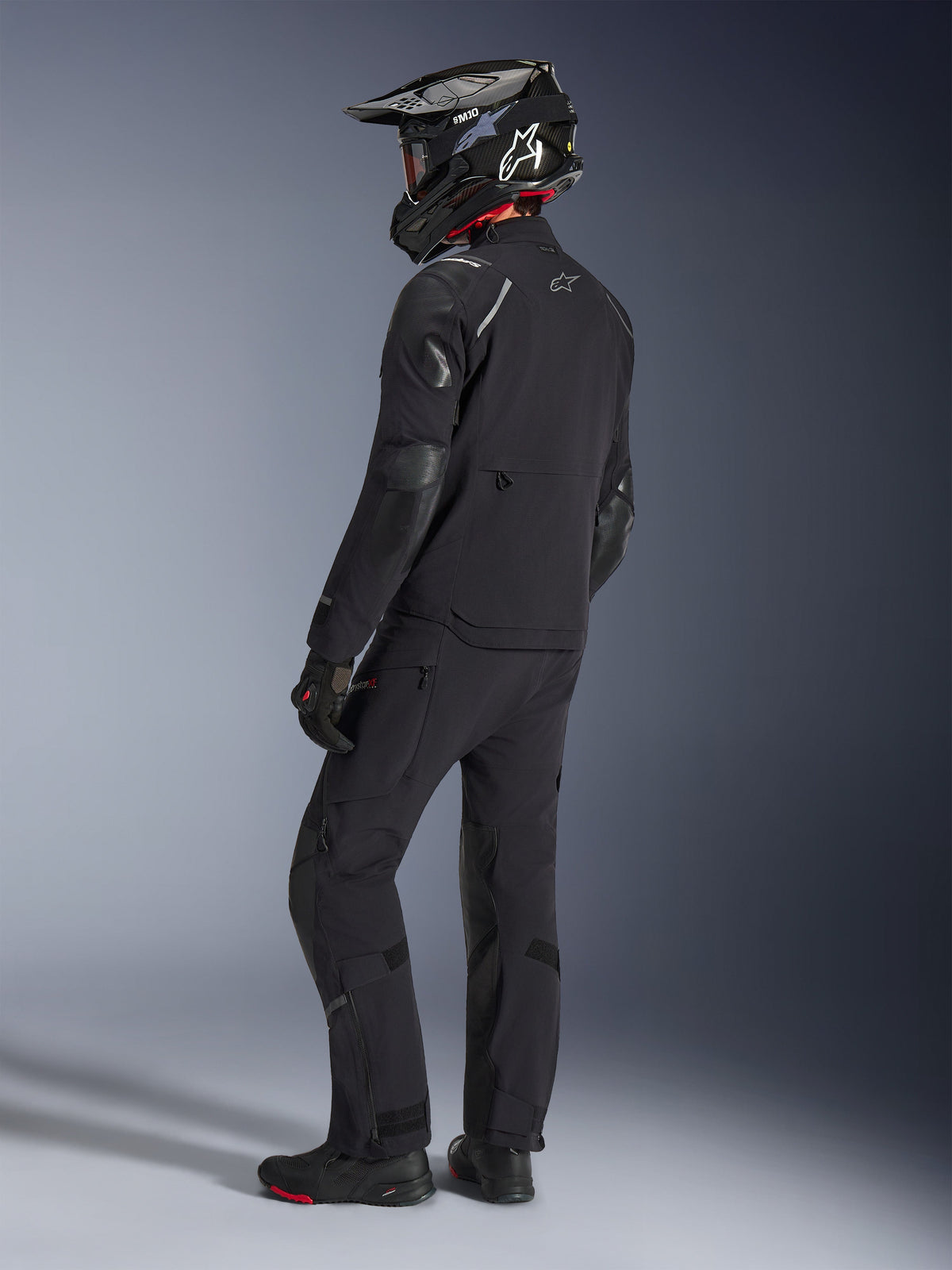 Motocyklista ubrany w kurtkę Alpinestars Andes Pro Drystar® XF i pasujące spodnie w kolorze czarnym i ciemnoszarym, widok z tyłu z profilu, z czarnym kaskiem adventure z włókna węglowego, czarnymi rękawicami i butami adventure, wysokiej klasy skoordynowany zestaw odzieży do turystyki przygodowej.