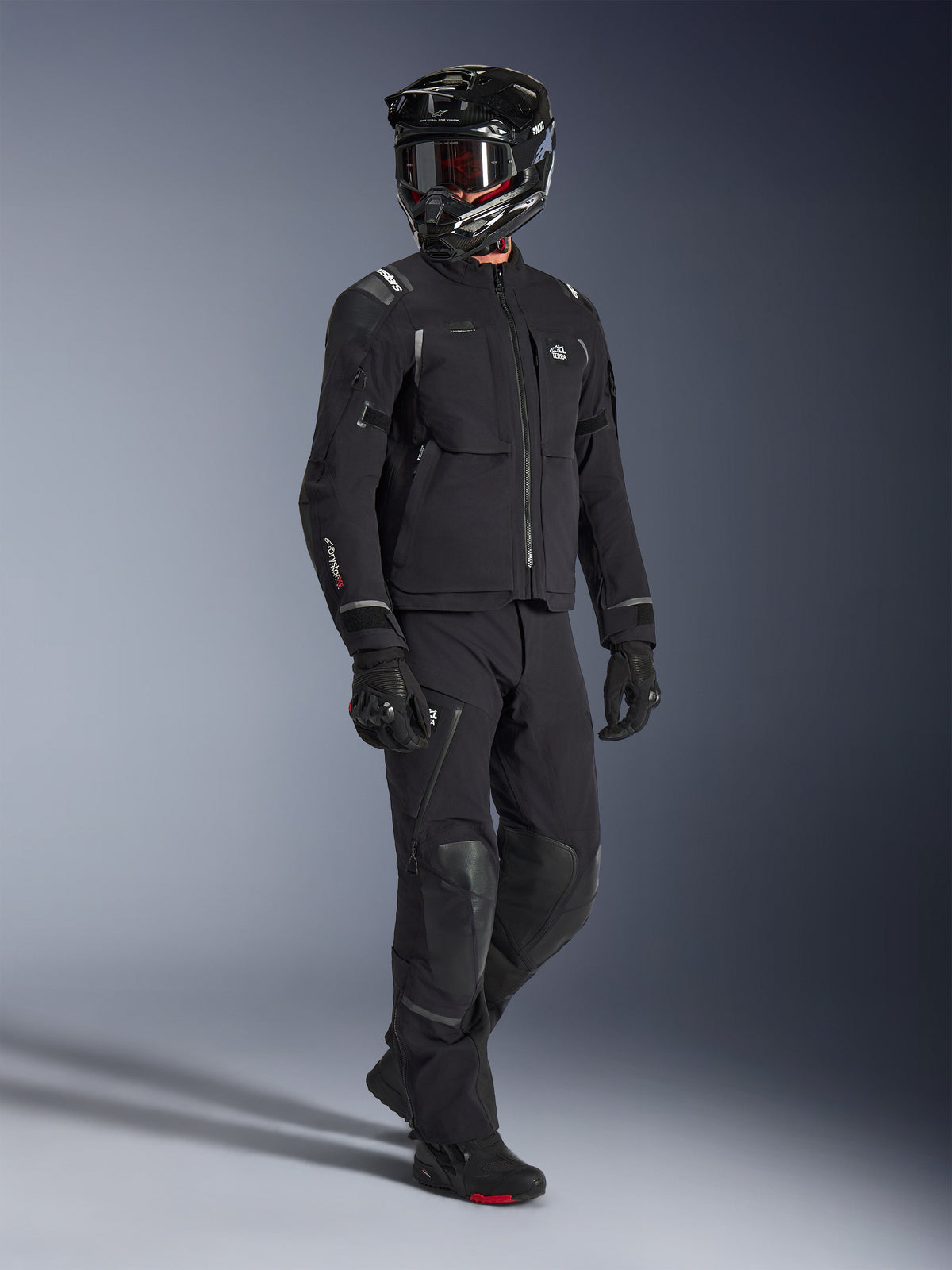 Kurtka Alpinestars Andes Pro Drystar® XF, czarno-ciemnoszara kurtka motocyklowa adventure, noszona z pasującymi czarnymi spodniami tekstylnymi, czarnym kaskiem integralnym z goglami, rękawicami i butami, wyposażona w 3-warstwową laminowaną powłokę i liczne kieszenie do całorocznej jazdy adventure.