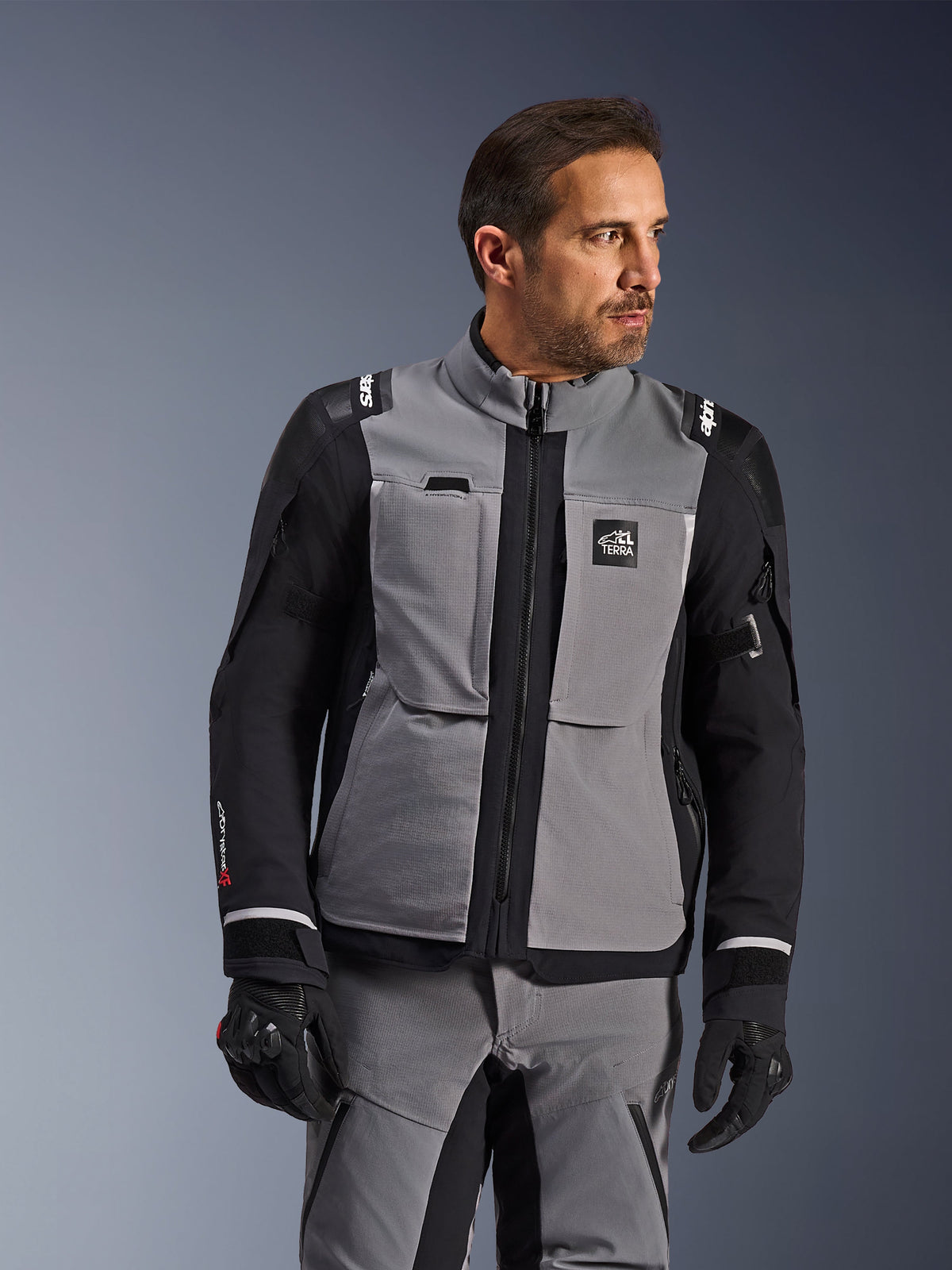 Alpinestars Andes Pro Drystar® XF Jacket in Dark Gray and Black, adventure riding gear, worn by a man with coordinated gray pants and black gloves, featuring a 3-layer laminated shell, utility pockets, and 'Terra' logo on the chest