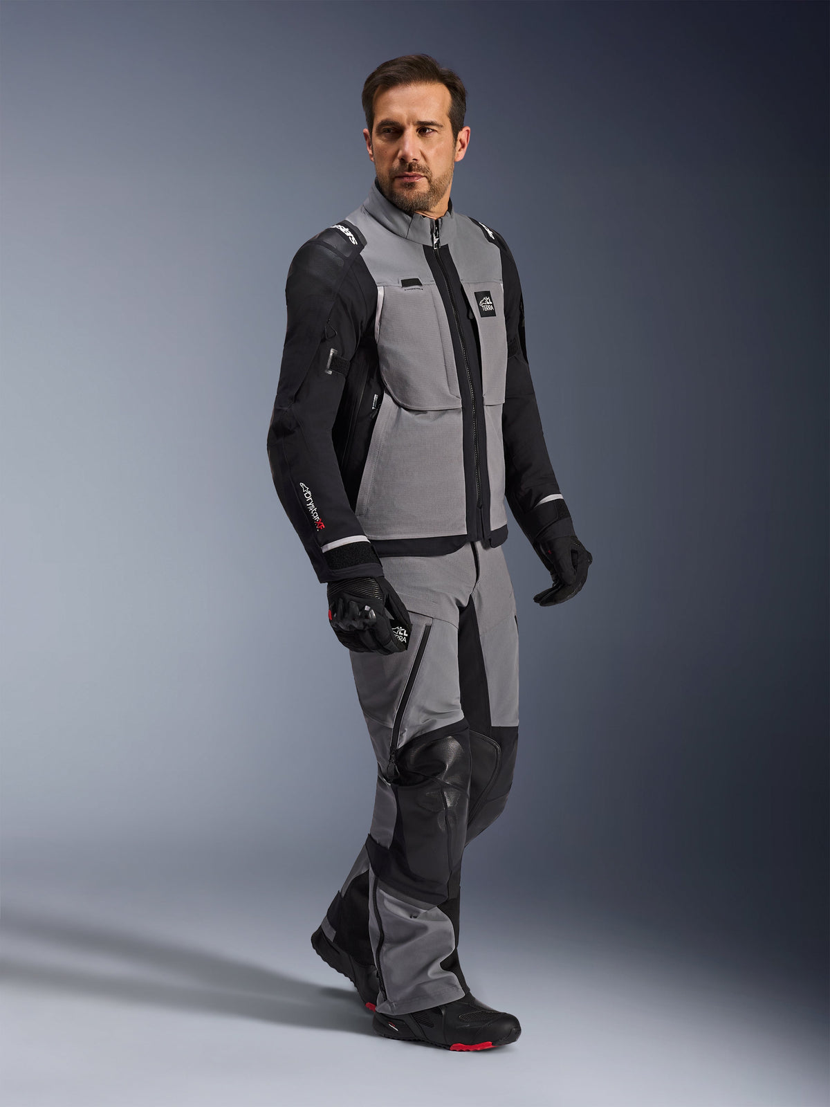 Alpinestars Andes Pro Drystar® XF Jacket, Adventure Motorcycle Jacket, Dark Gray and Black, worn by a model with matching textile pants and black gloves, featuring a multi-pocket design, technical 3-layer laminated shell, and articulated fit for long-distance adventure riding.