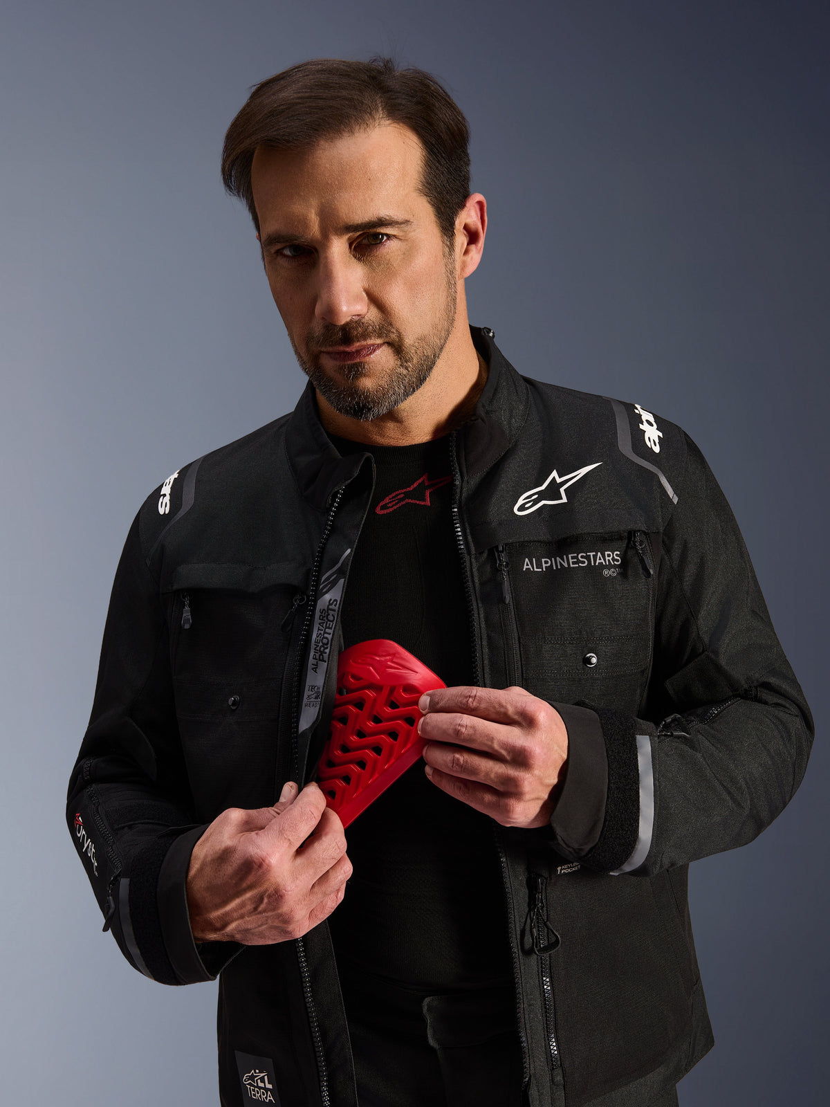 Alpinestars Cusco Drystar®, czarna kurtka motocyklowa adventure, model trzymający czerwony ochraniacz Nucleon Flex Pro.