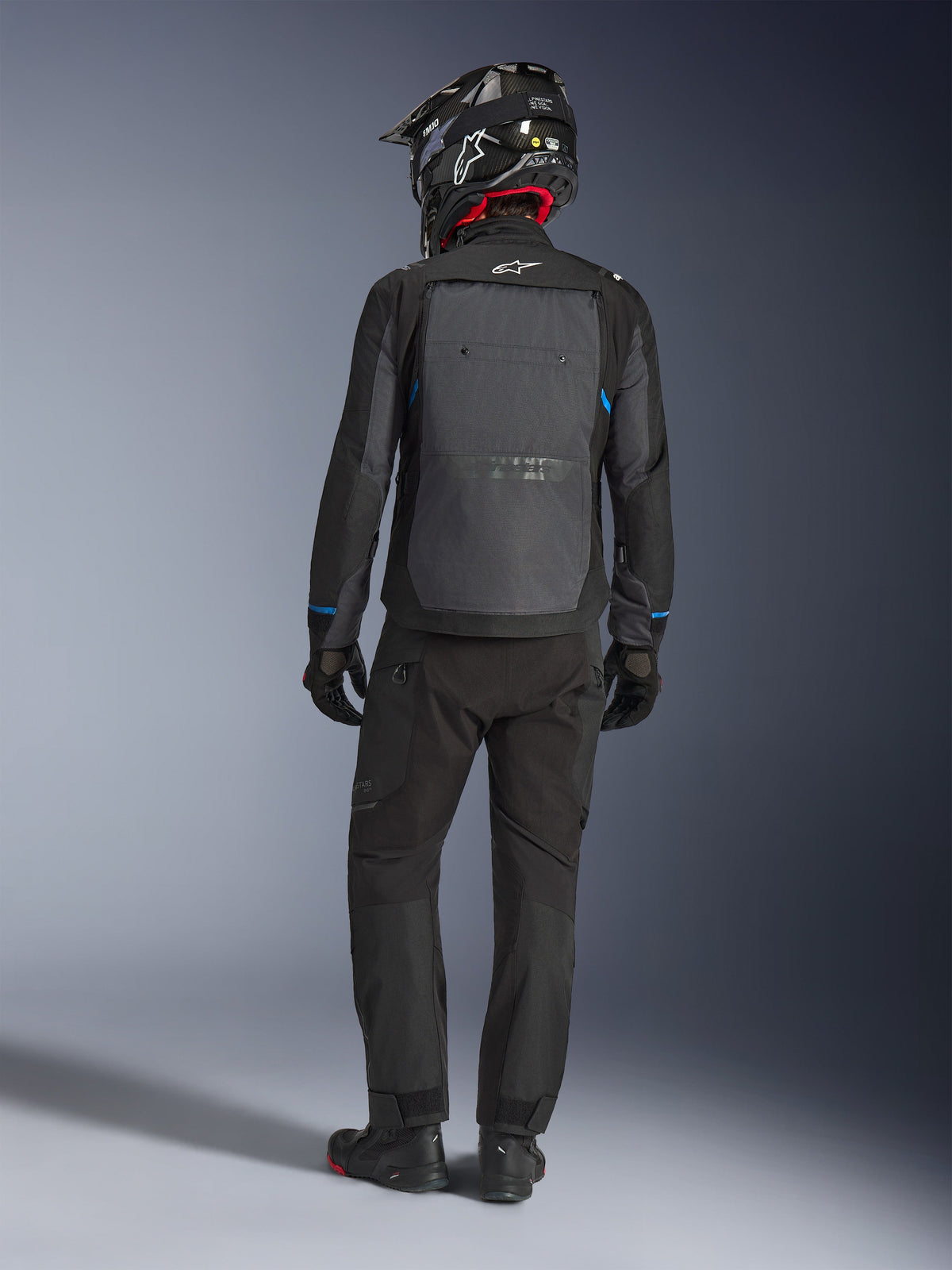 Kurtka Alpinestars Cusco Drystar®, czarna i ciemnoszara, widok z tyłu motocyklisty w pełnym stroju adventure touring, duży szary panel na plecach z logo Alpinestars, niebieskie akcenty na rękawach, noszona z pasującymi czarnymi spodniami i kaskiem.