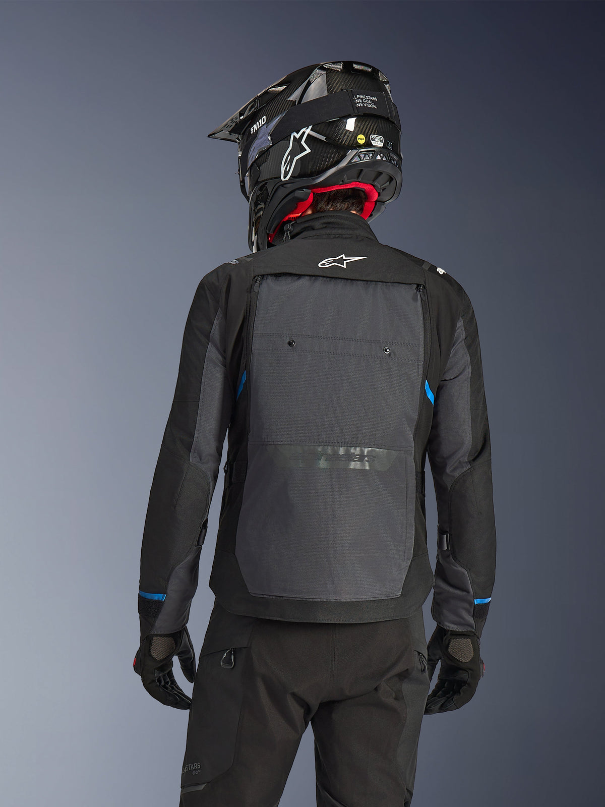 Motocyklista widziany od tyłu w kurtce Alpinestars Cusco Drystar® w kolorze czarnym i ciemnoszarym, z dużym szarym panelem wentylacyjnym na plecach i białym logo, w czarnym kasku terenowym i czarnym stroju do jazdy adventure touring.
