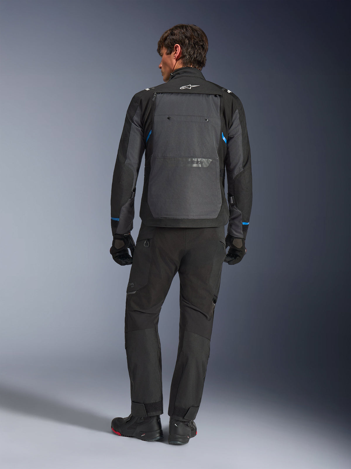Alpinestars Cusco Drystar® Jacket, czarno-ciemnoszara z niebieskimi akcentami, widok z tyłu, noszona z dopasowanymi czarnymi spodniami adventure i rękawicami, z dużym szarym panelem na plecach i białym logo na kołnierzu, zaprojektowana do turystyki szosowej i adventure touring