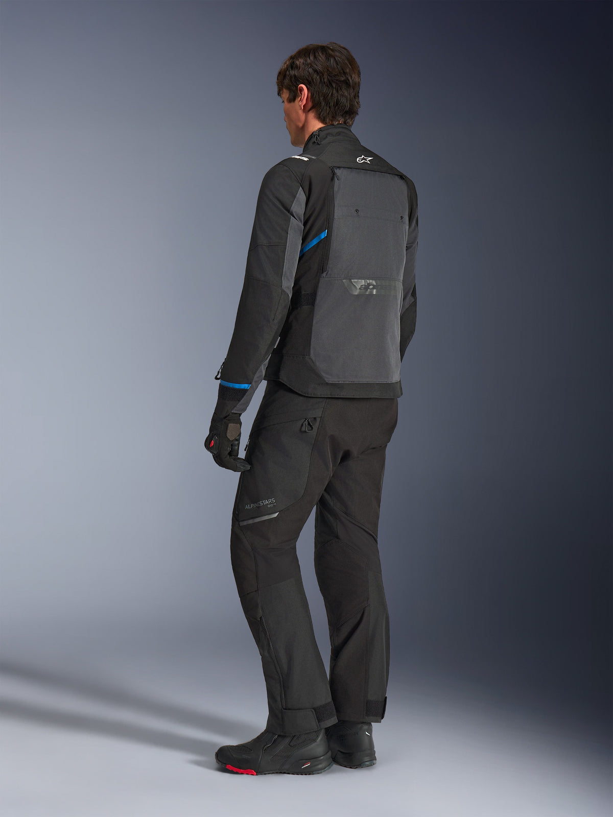 Kurtka Alpinestars Cusco Drystar®, czarna i ciemnoszara, widok z tyłu z boku prezentujący duży tylny panel wentylacyjny oraz niebieskie akcenty na rękawach, noszona z czarnymi spodniami motocyklowymi i butami dla kompletnego stroju turystycznego.