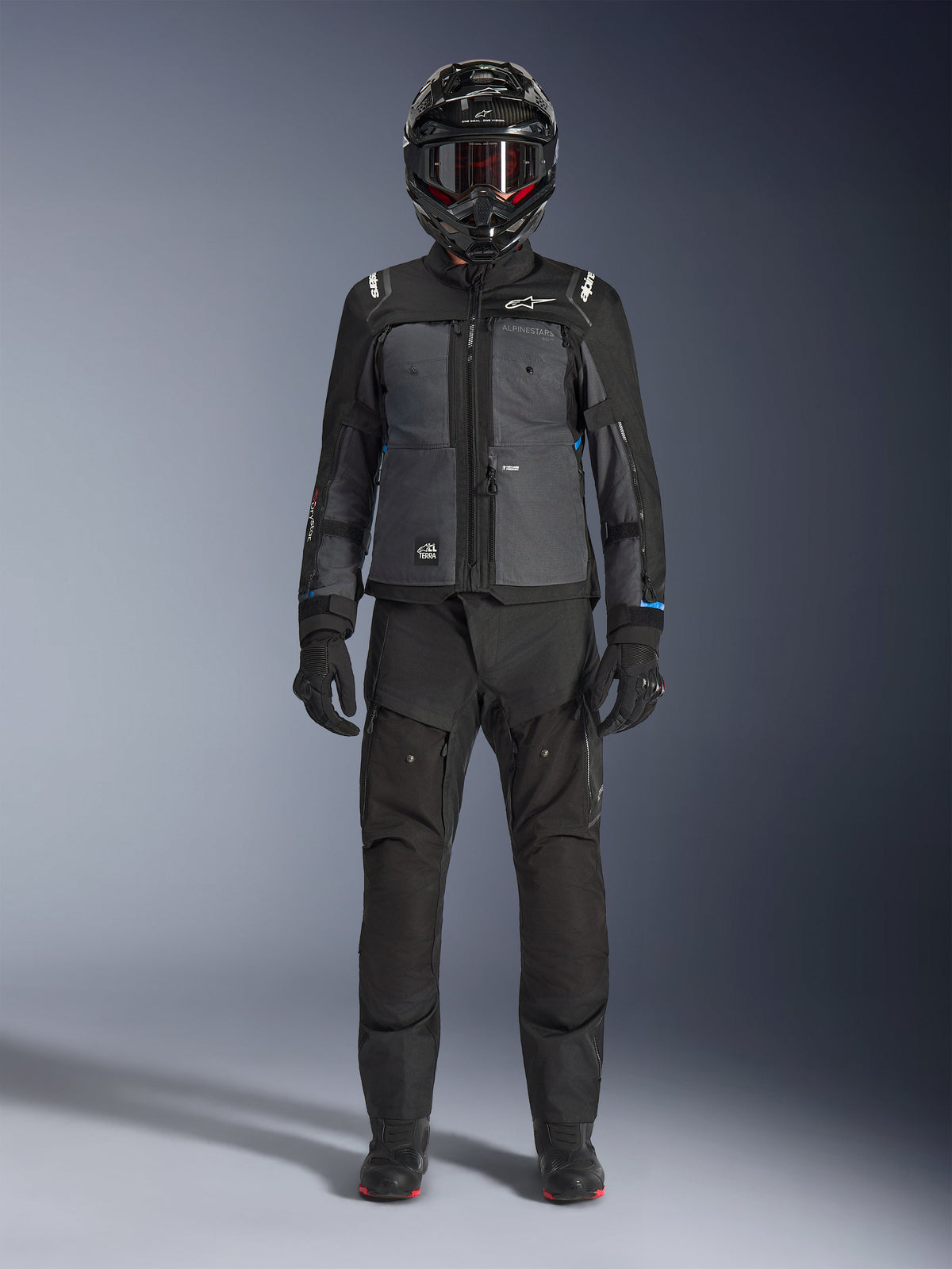 Kurtka Alpinestars Cusco Drystar®, czarno-ciemnoszara kurtka adventure touring, noszona z dopasowanymi czarnymi spodniami, rękawicami i kaskiem integralnym, widok na całą sylwetkę od przodu, ochronna odzież motocyklowa