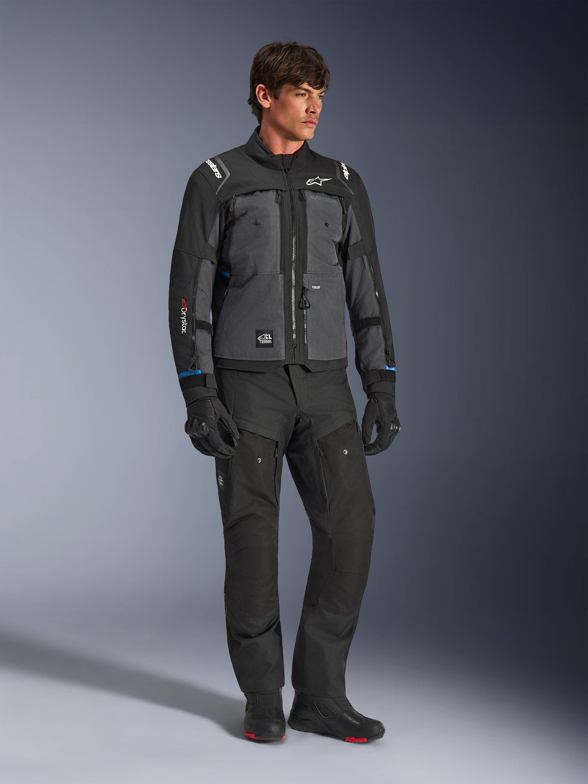 Kurtka Alpinestars Cusco Drystar® w kolorze czarnym i ciemnoszarym, techniczna motocyklowa kurtka turystyczna z wielotonowymi panelami, noszona z pasującymi czarnymi spodniami i rękawicami, zaprojektowana do jazdy szosowej i turystyki adventure.