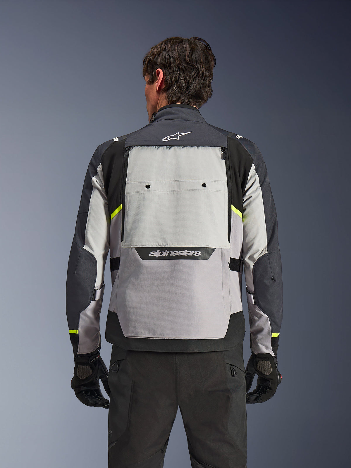 Widok od tyłu motocyklisty w kurtce Alpinestars Cusco Drystar® w kolorach Ice Gray, Mid Gray i Deep Gray, z neonowożółtymi akcentami, dużym panelem wentylacyjnym na plecach i wyraźnym logo Alpinestars, w zestawieniu z czarnymi spodniami i rękawicami motocyklowymi.