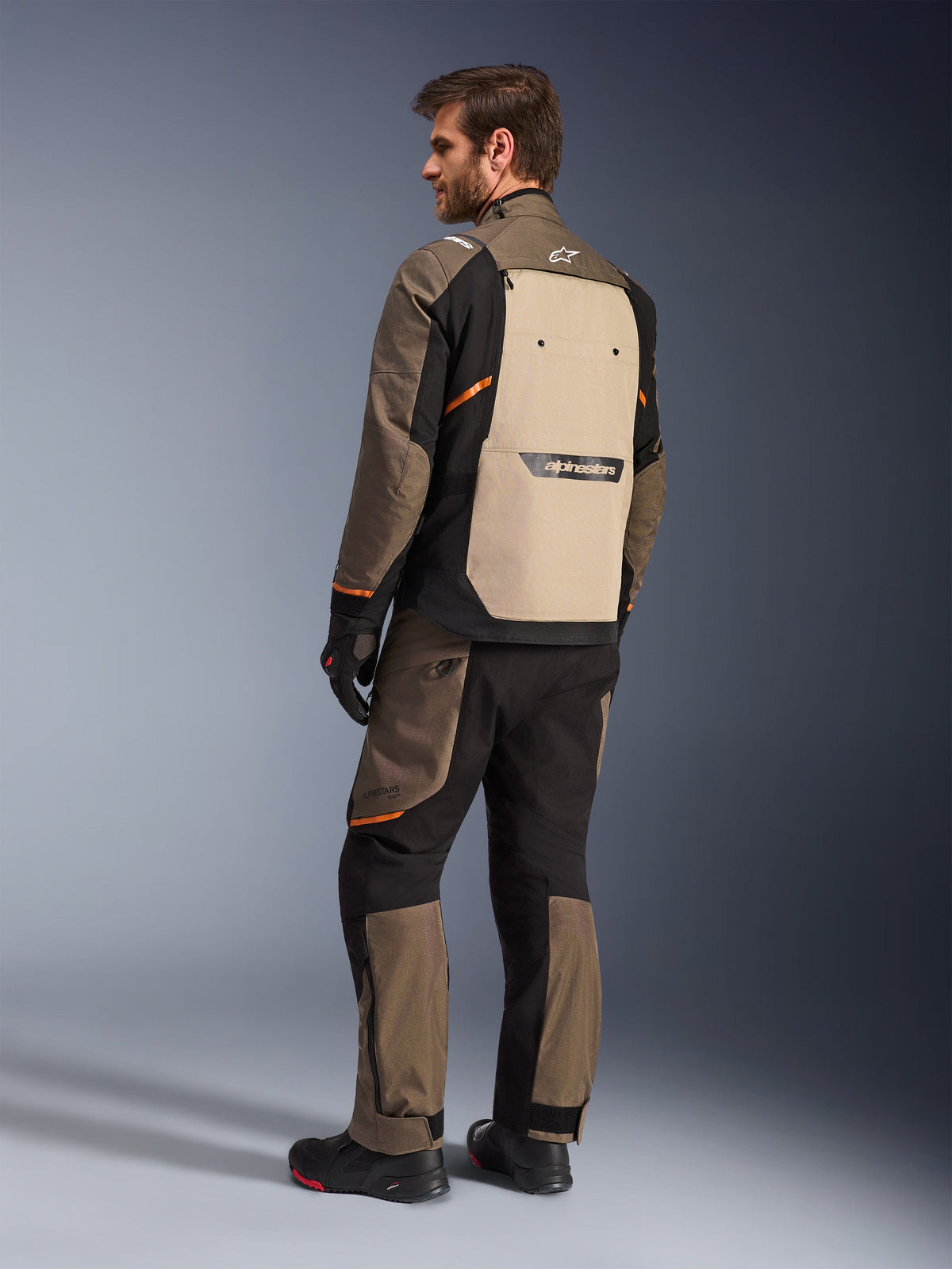 Kurtka Alpinestars Cusco Drystar® w kolorze Walnut Aluminum Black, widok z tyłu ukazujący beżowy panel tylny i pomarańczowe akcenty, noszona z pasującymi spodniami motocyklowymi, czarnymi rękawicami i butami, profesjonalna odzież adventure touring