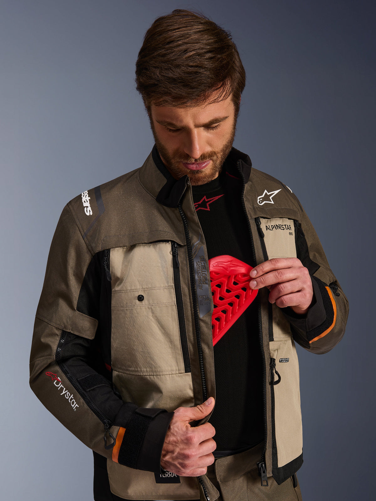 Kurtka Alpinestars Cusco Drystar®, Walnut Aluminum Black, noszona przez mężczyznę prezentującego kieszeń na ochraniacz klatki piersiowej z czerwoną wkładką ochronną, tekstylna kurtka motocyklowa z otworami wentylacyjnymi na zamek i brandingiem adventure touring.