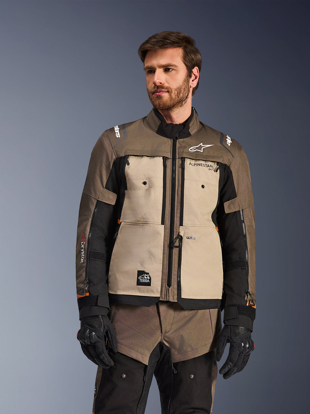 Kurtka Alpinestars Cusco Drystar®, przygodowa kurtka motocyklowa, kolor Walnut Aluminum Black, prezentowana przez modela w czarnych rękawicach, z beżowymi panelami na klatce piersiowej i czarnymi rękawami, logo Alpinestars i naszywka All Terra, ochronna odzież turystyczna na każdą pogodę
