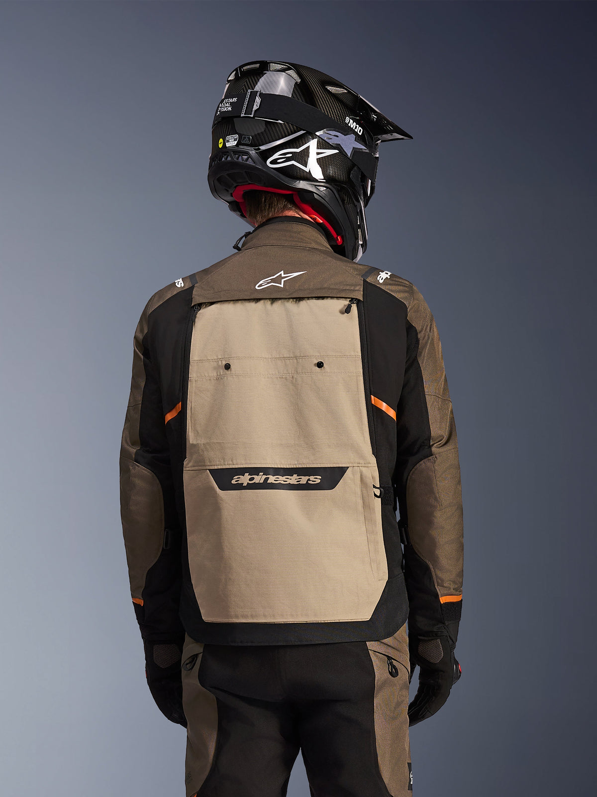 Widok z tyłu kurtki Alpinestars Cusco Drystar® w kolorze Walnut Aluminum Black, motocyklowa kurtka adventure touring z dużym beżowym panelem na plecach z logo Alpinestars, czarnymi rękawami z pomarańczowymi akcentami, pokazana z czarnym kaskiem w stylu karbonowym.