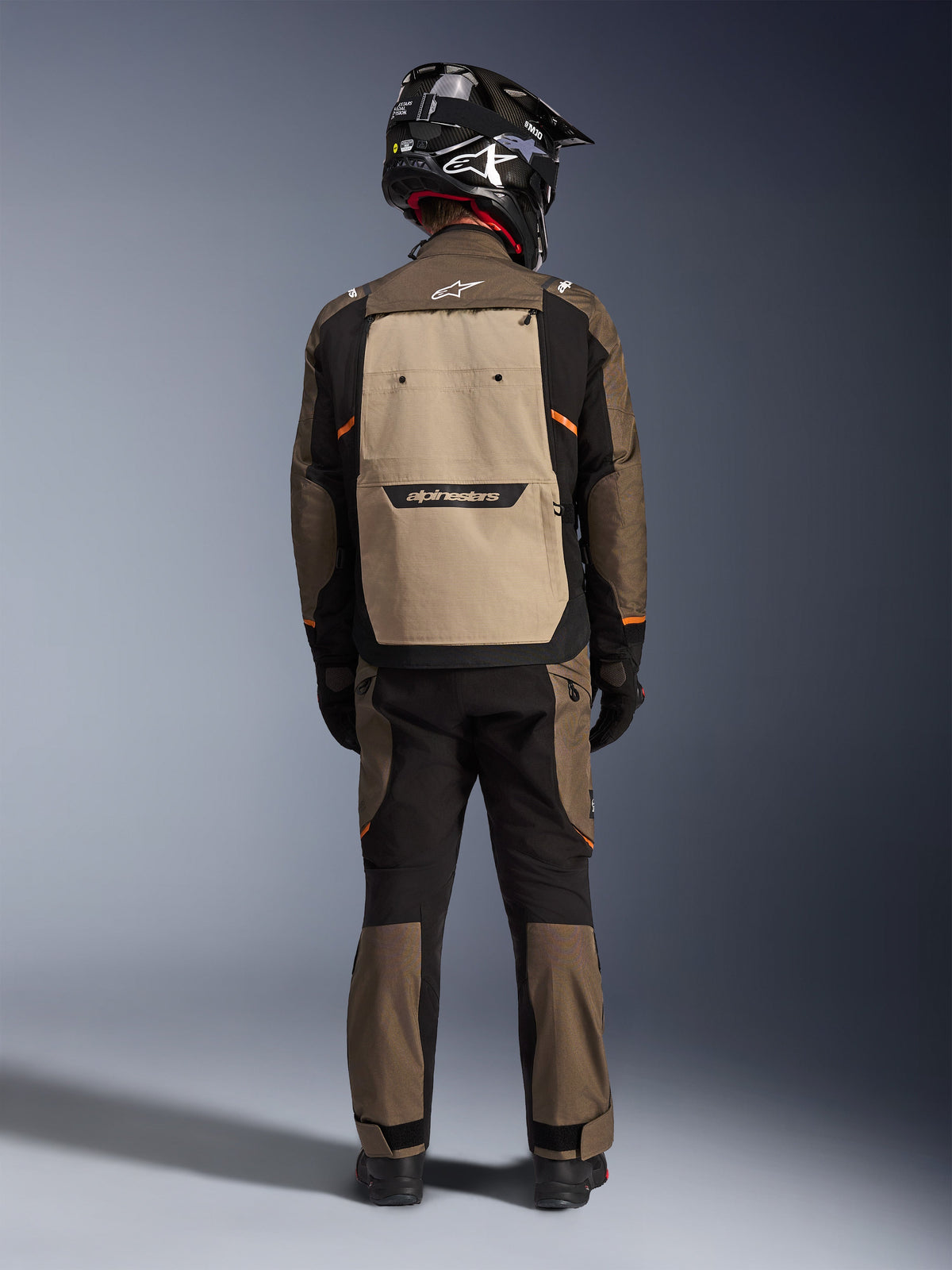 Motocyklista widziany od tyłu w kurtce Alpinestars Cusco Drystar® w kolorze Walnut Aluminum Black, z dużą tylną kieszenią w kolorze aluminium i pomarańczowymi akcentami, pasujące spodnie adventure, czarny kask off-road i rękawice, wytrzymała tekstylna odzież motocyklowa turystyczna.