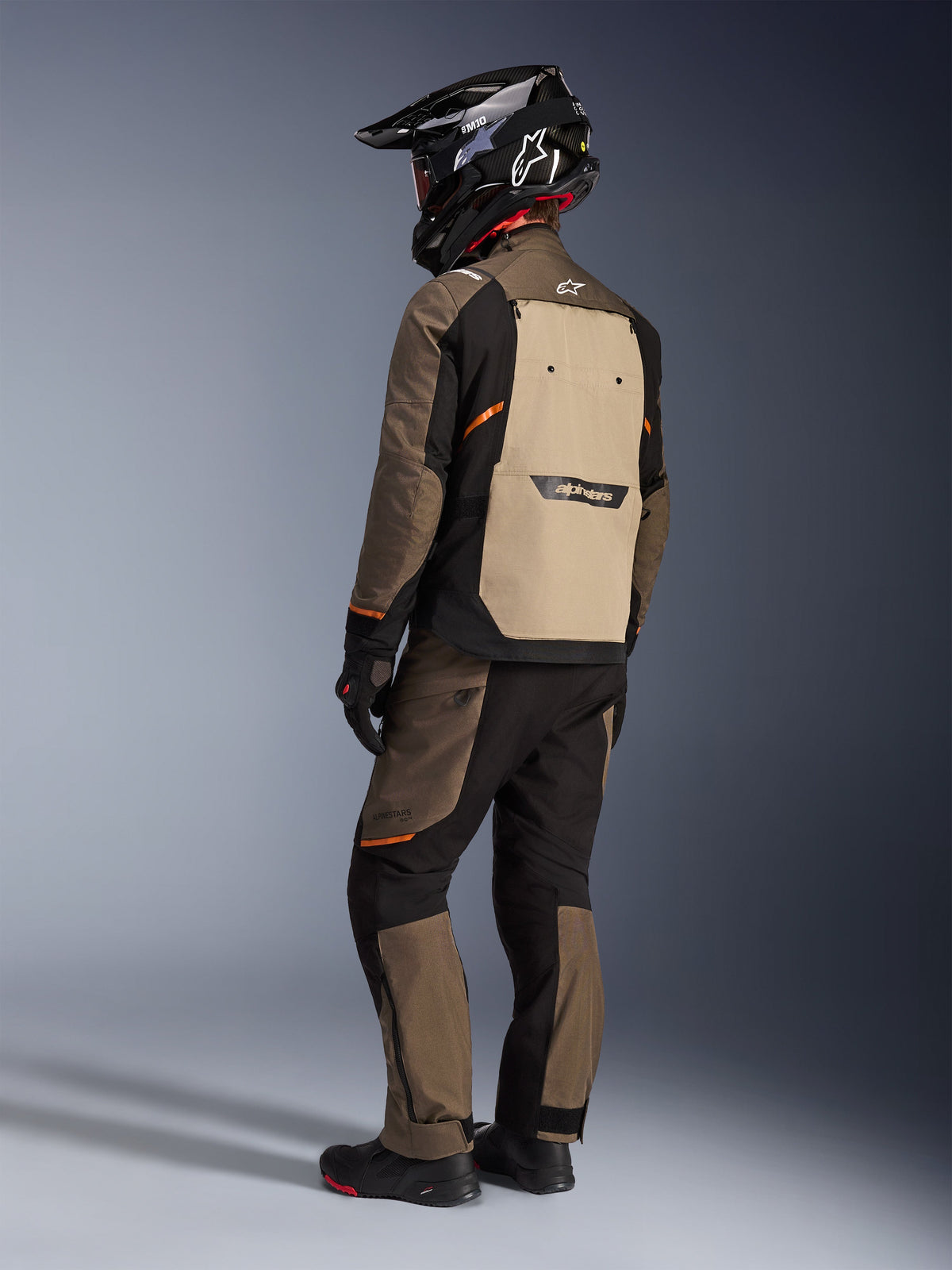 Kurtka Alpinestars Cusco Drystar®, kurtka turystyczna adventure, Walnut Aluminum Black, widok z tyłu prezentujący beżowy panel tylny i pomarańczowe akcenty, noszona z pasującymi spodniami i czarnym kaskiem z włókna węglowego