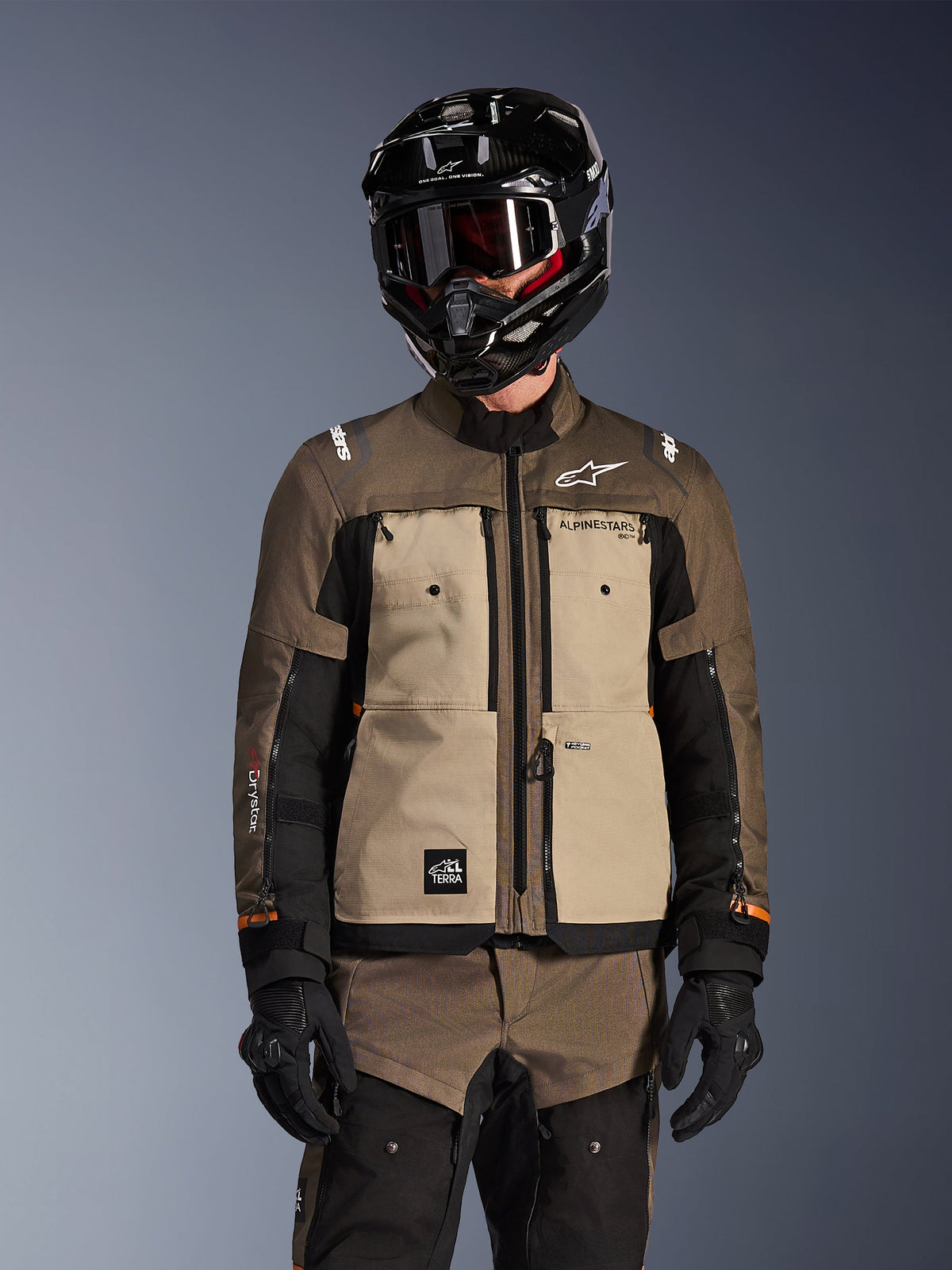 Motocyklista w kurtce Alpinestars Cusco Drystar w kolorze Walnut Aluminum Black, motocyklowy strój typu adventure touring, czarny kask z goglami i czarne rękawice, widok z przodu przedstawiający techniczną konstrukcję tekstylną, kieszenie oraz branding.