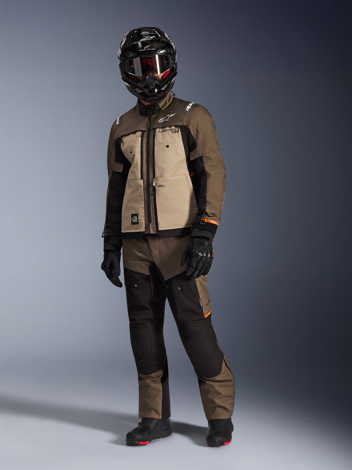 Kurtka Alpinestars Cusco Drystar®, Walnut Aluminum Black, noszona przez motocyklistę w pełnym stroju adventure touring, piaskowe i brązowe panele z czarnymi akcentami, dopasowane spodnie, czarny kask z goglami i czarne buty, tło studyjne