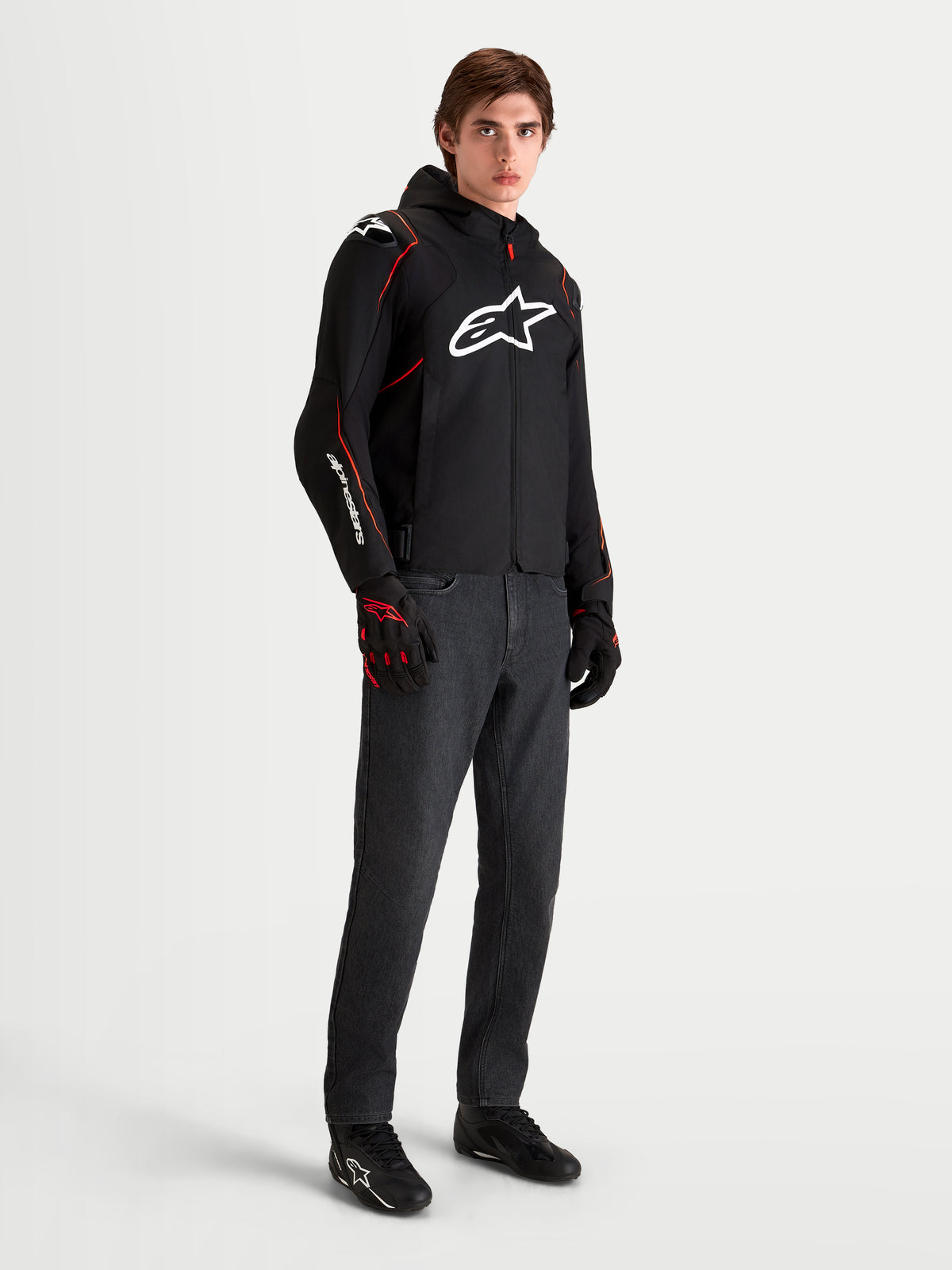 Alpinestars Zaca V2 Waterproof, męska czarno-czerwona motocyklowa kurtka sportowa z kapturem i białym logo na piersi, prezentowana przez modela w pasujących rękawicach i ciemnych jeansach