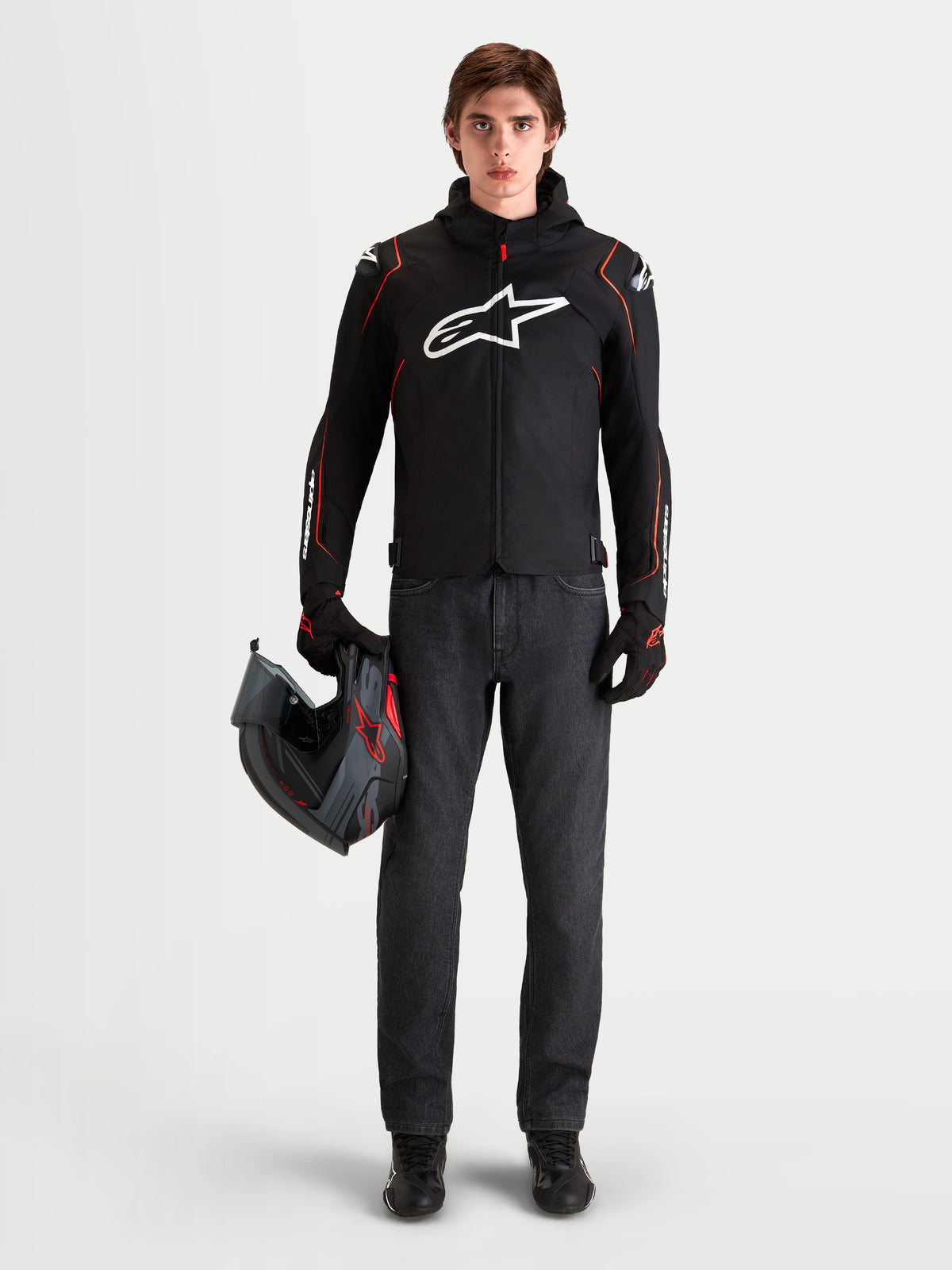 Kurtka Alpinestars Zaca V2 Waterproof, męska czarno-czerwona motocyklowa kurtka sportowa z ochraniaczami ramion DFS i zintegrowanym kapturem, prezentowana przez modela trzymającego kask
