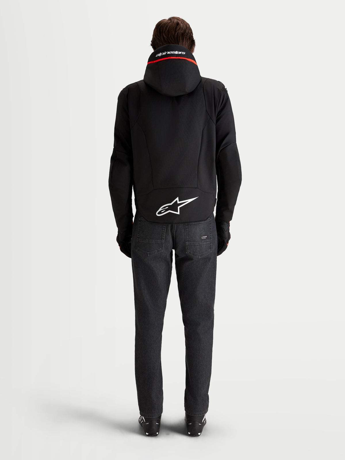 Alpinestars Zaca V2 Waterproof kurtka motocyklowa męska, czarna zintegrowany kaptur, widok z tyłu z przedłużonym dołem i białe logo Astar, ciemne jeansy
