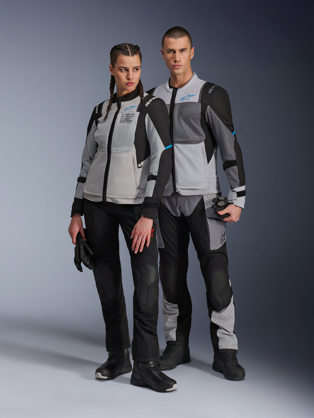 Mężczyzna i kobieta w kurtkach Alpinestars Stella ST-2 Air i pasujących spodniach technicznych, kolorystyka Ice Gray i Black z niebieskimi akcentami, panele wentylacyjne z siatki, branding „ALPINESTARS EXPLORATION EQUIPMENT”, studyjne ujęcie pary