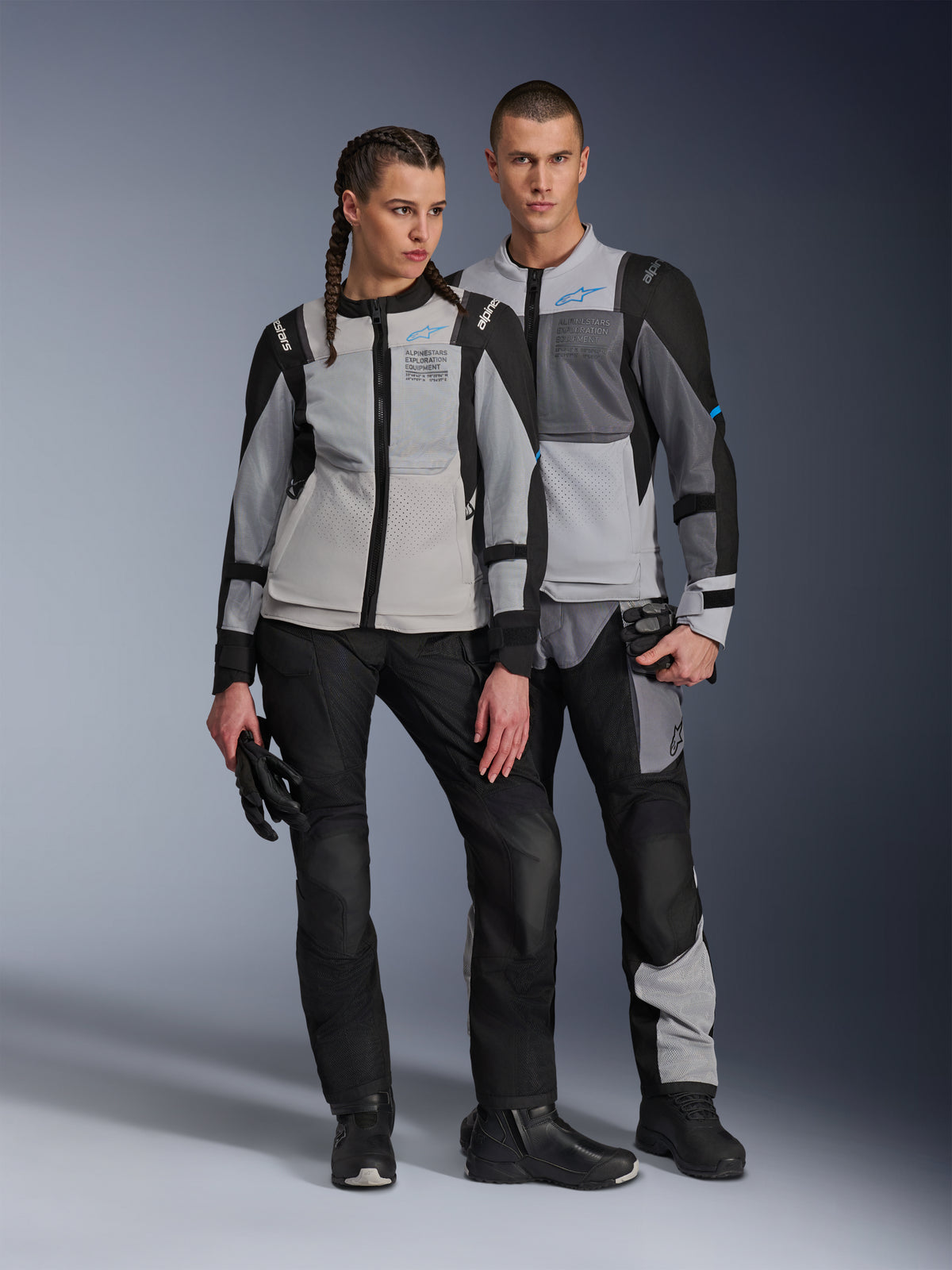 Modele (kobieta i mężczyzna) ubrani w kurtki Alpinestars ST-2 Air w kolorach Ice Gray, Dark Gray i Black, techniczna letnia turystyczna odzież motocyklowa z dużymi panelami z siatki i brandingiem exploration equipment, w zestawie z czarnymi spodniami, rękawicami i butami.