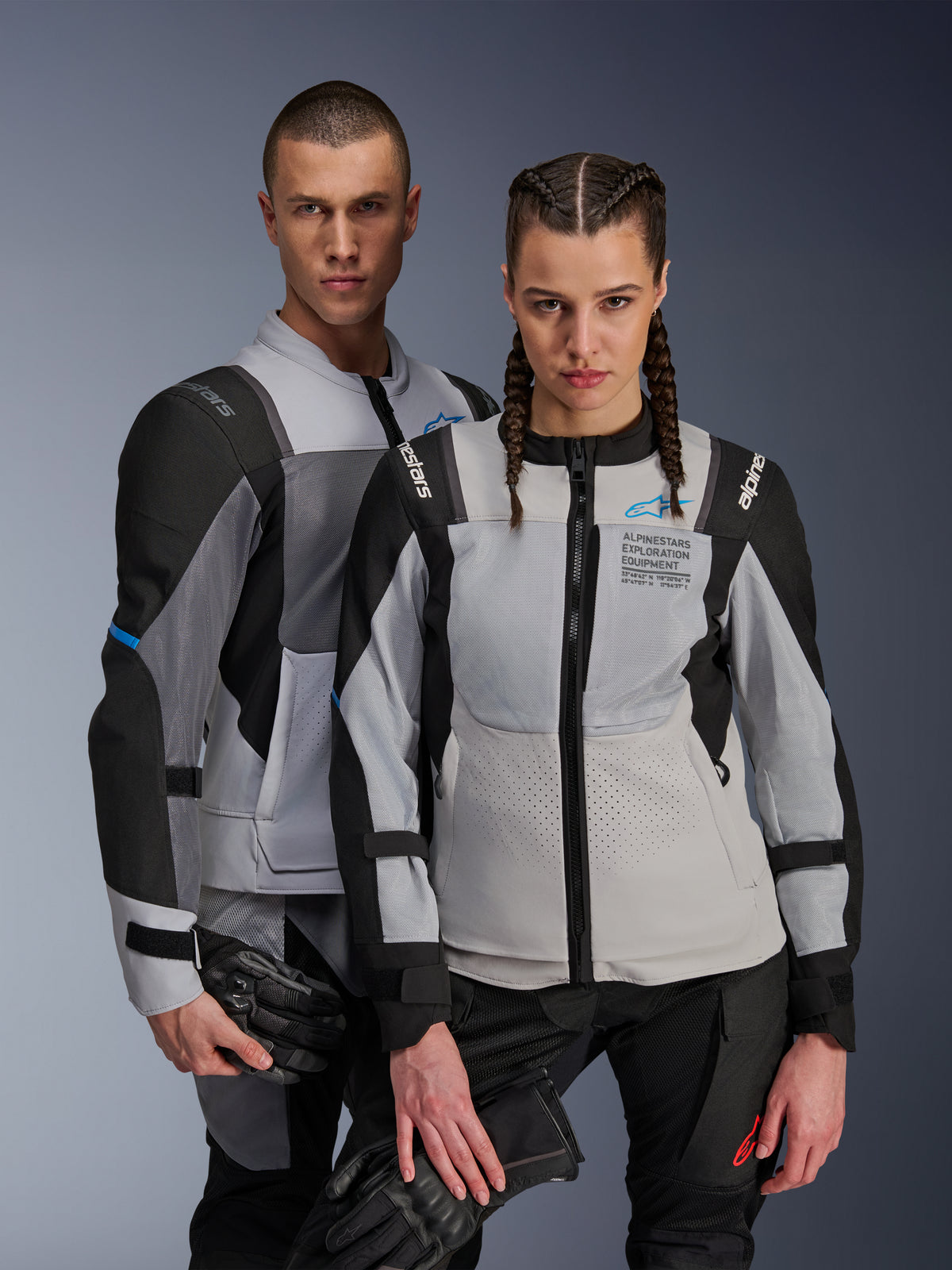 Kurtki Alpinestars ST-2 Air i Stella ST-2 Air, kolor lodowo-szary i czarny, tekstylne kurtki motocyklowe z panelami z siatki i laserowymi perforacjami, z brandingiem „Alpinestars Exploration Equipment”, noszone przez modela i modelkę trzymających rękawice motocyklowe.