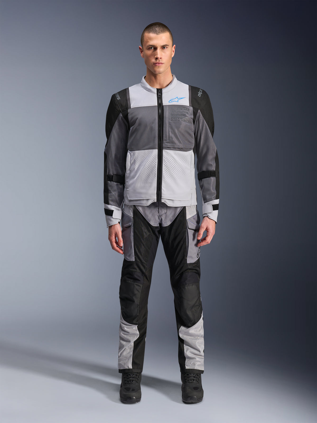 Kurtka Alpinestars ST-2 Air, motocyklowa kurtka turystyczna, kolory lodowy szary, ciemnoszary i czarny, prezentowana na modelu z dopasowanymi spodniami adventure, duże panele z siatki, niebieskie logo Alpinestars na klatce piersiowej, branding Alpinestars Exploration Equipment