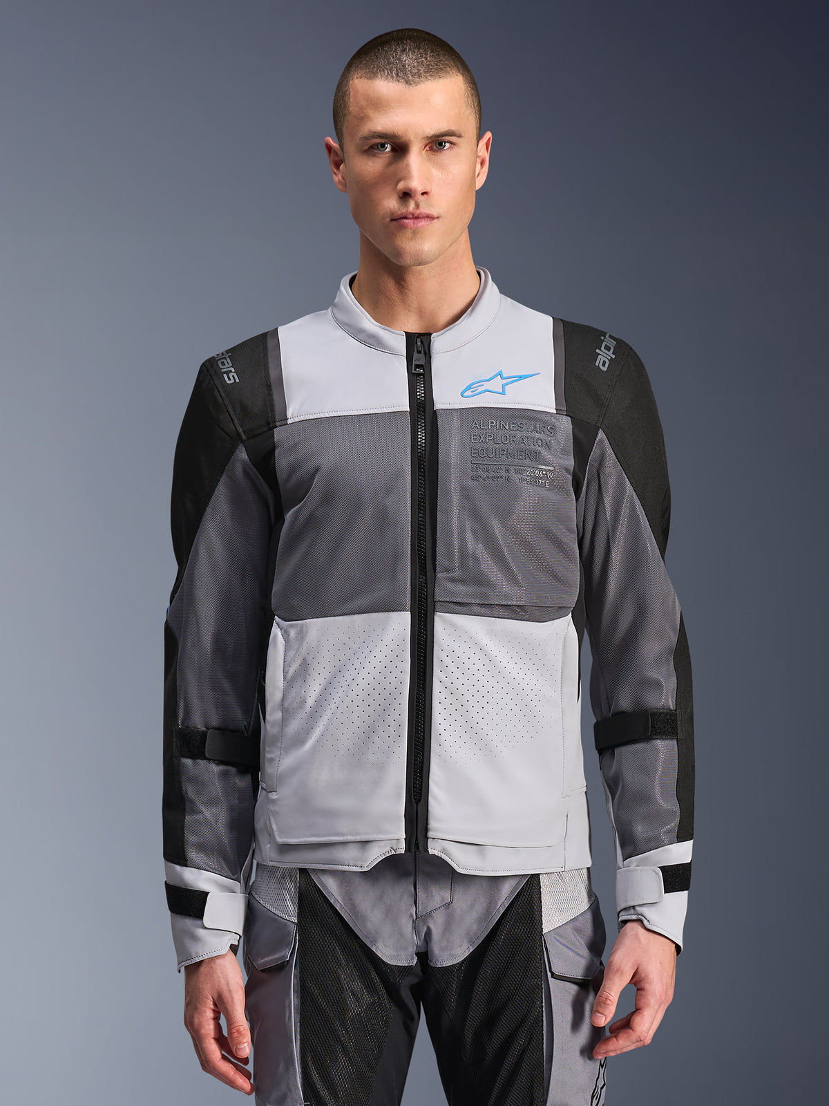 Kurtka Alpinestars ST-2 Air, męska kurtka motocyklowa, jasnoszara i ciemnoszara z czarnymi akcentami, szerokie panele wentylacyjne z siatki, laserowo perforowany przód, niebieskie logo Alpinestars, przedni zamek, prezentowana przez modela z pasującymi spodniami do jazdy