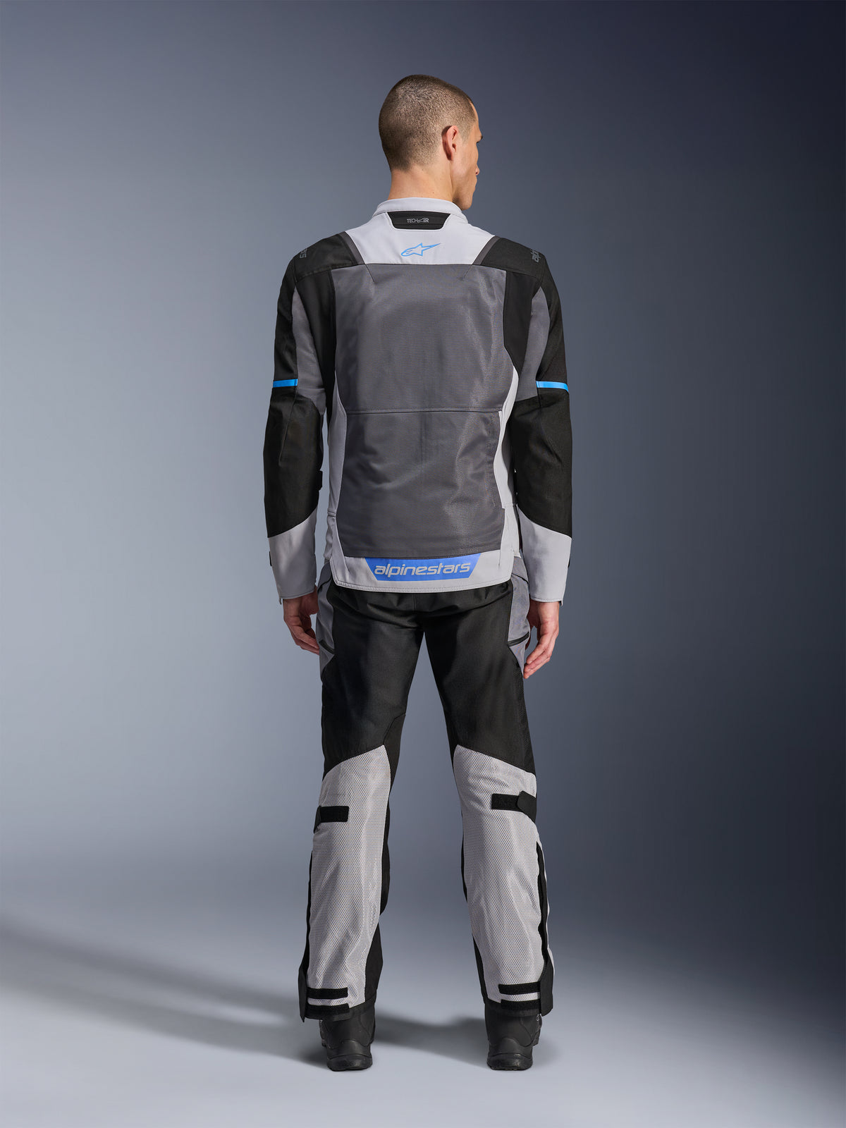 Alpinestars ST-2 Air Jacket, kolor Ice Gray Dark Gray Black, widok z tyłu na modelu, duże siatkowe panele wentylacyjne, niebieskie akcenty i logo Alpinestars, w zestawie z pasującymi spodniami motocyklowymi do adventure touring.