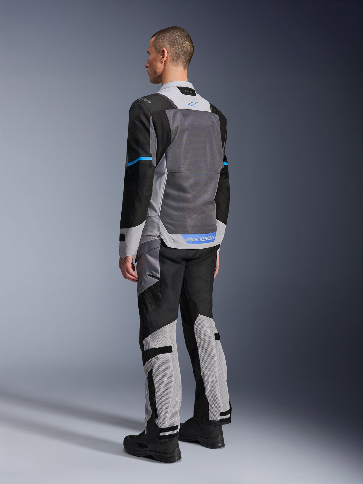 Męski model widoczny od tyłu w kurtce Alpinestars ST-2 Air w kolorach Ice Gray, Dark Gray i czarnym, z dużym szarym panelem z siatki na plecach, niebieskimi akcentami logo i dopasowanymi spodniami motocyklowymi do letniej turystyki.