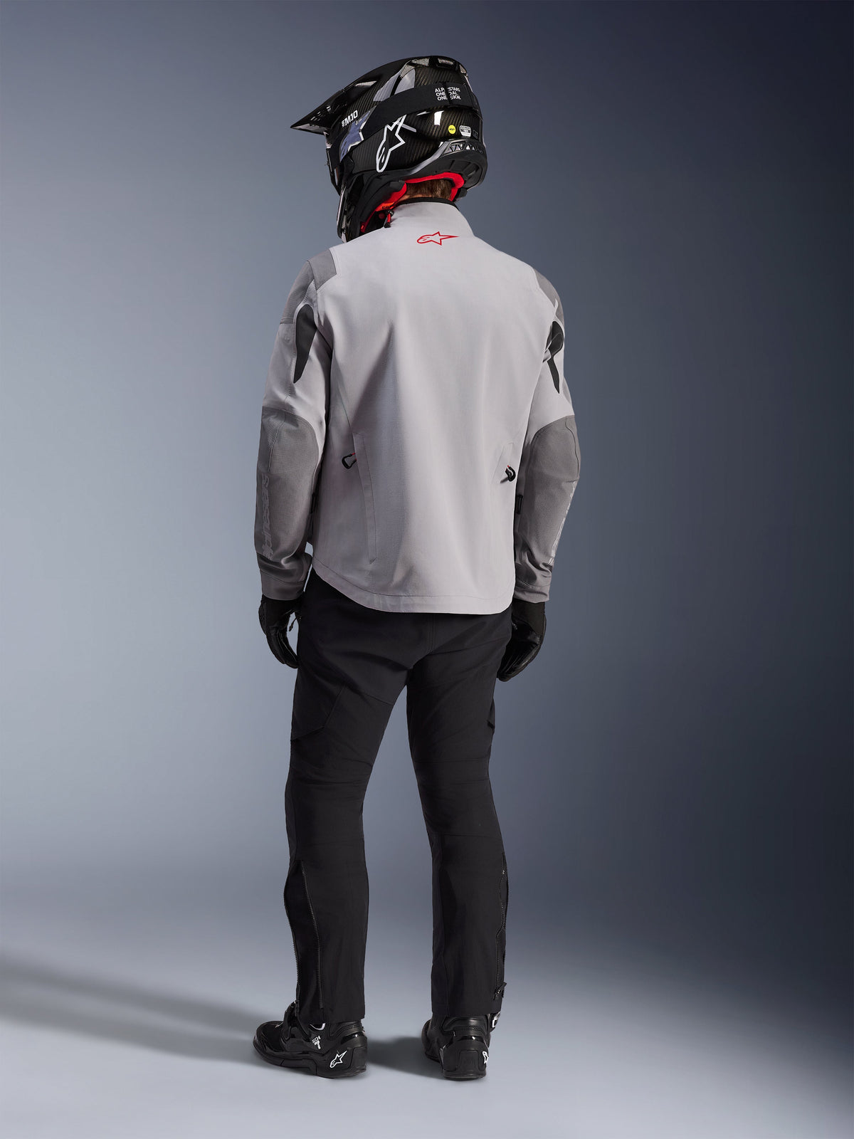 Widok z tyłu motocyklisty ubranego w kurtkę Alpinestars Maxdura Drystar®XF Laminated Armored Shell w kolorach Mid Gray i Dark Gray, z czerwonym logo gwiazdy na kołnierzu, w zestawie z czarnymi spodniami adventure, czarnym kaskiem z karbonowym wzorem i czarnymi butami.
