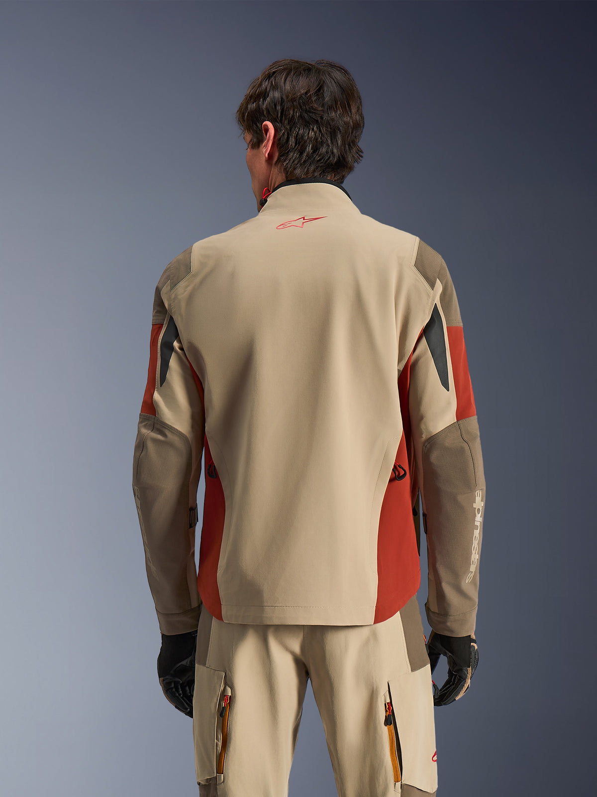 Mężczyzna w kurtce Alpinestars Maxdura Drystar®XF Laminated Armored Shell widoczny od tyłu, kurtka adventure w kolorze Aluminum Walnut i Rust Brown, pasujące spodnie techniczne i czarne rękawice, 3-warstwowa laminowana konstrukcja tekstylna ze wzmocnionymi panelami, dopasowany strój do turystyki motocyklowej