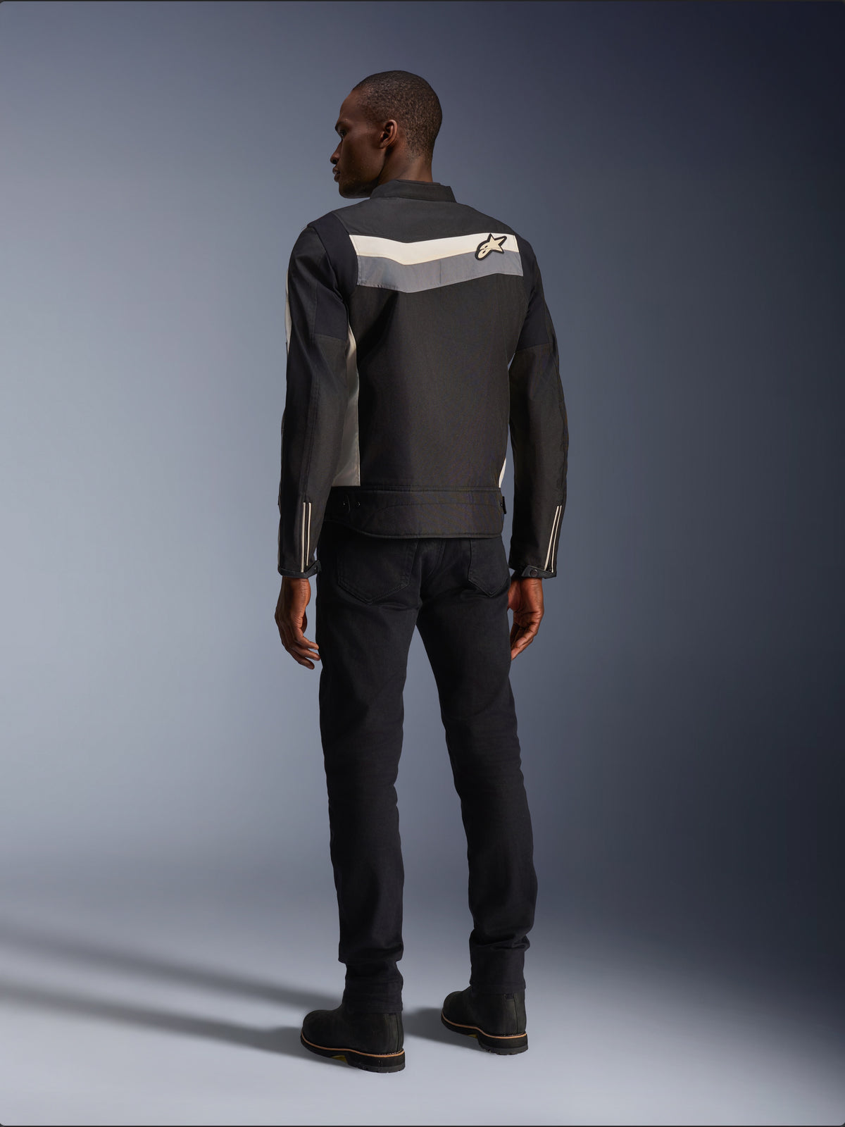 Male model from behind wearing the Alpinestars T-Dyno WR Jacket in Black Dark Gray Ecru, textile motorcycle jacket with a horizontal stripe and star logo on the back, paired with black riding pants and boots, urban motorcycle gear aesthetic.