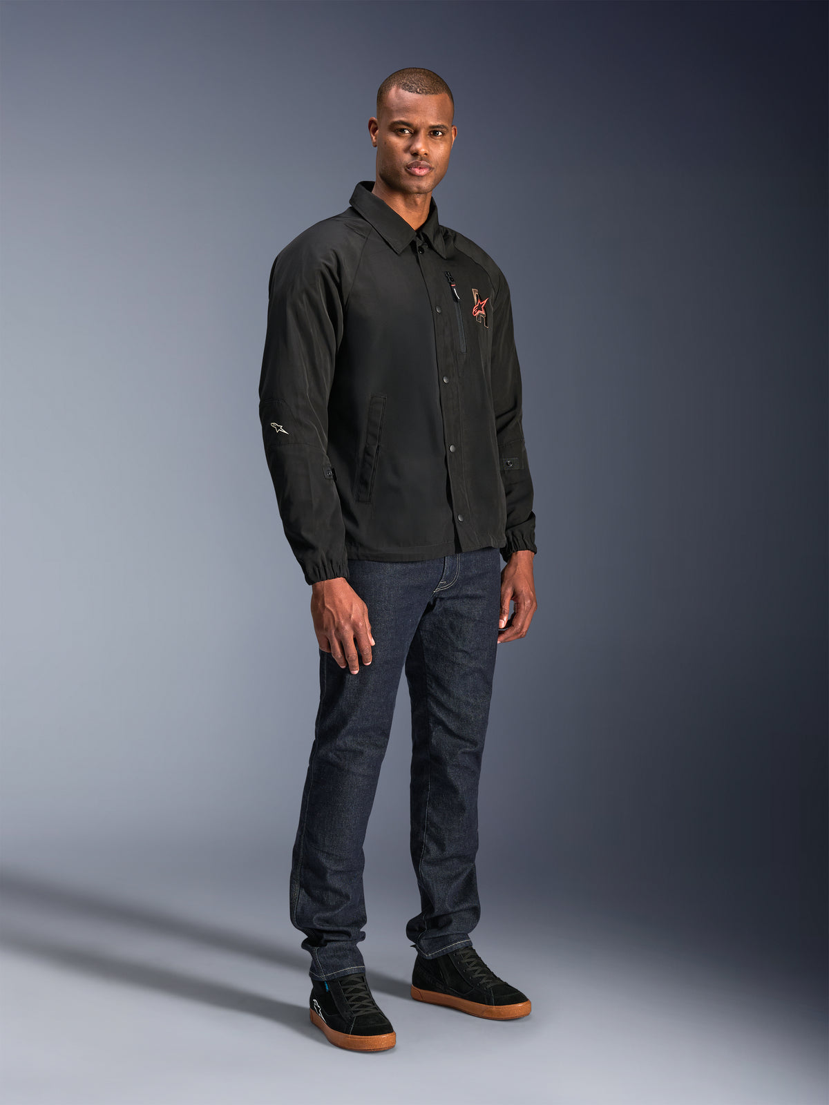 Alpinestars Revved WR Jacket in Black, urban coaches-style motorcycle jacket worn by a model with dark blue jeans and black sneakers, featuring a snap-button front and embroidered logo, minimalist and sporty design for road riding.