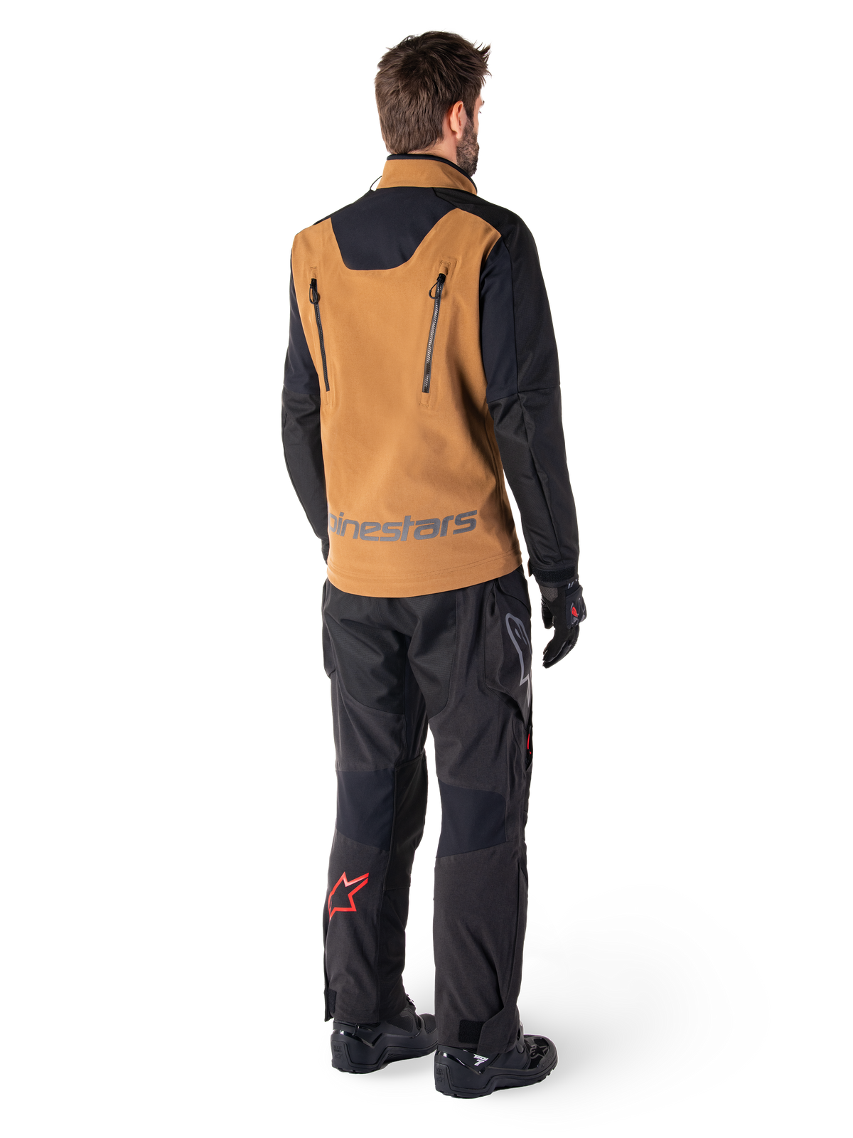 Kurtka Alpinestars Hyde XT Stretch Drystar®XF i spodnie Hyde XT Drystar®, widok z tyłu motocyklisty w beżowo-czarnym stroju adventure touring, kurtka z pionowymi zamkami na plecach i logo Alpinestars, czarne spodnie z czerwoną gwiazdą na łydce, czarne rękawice i buty