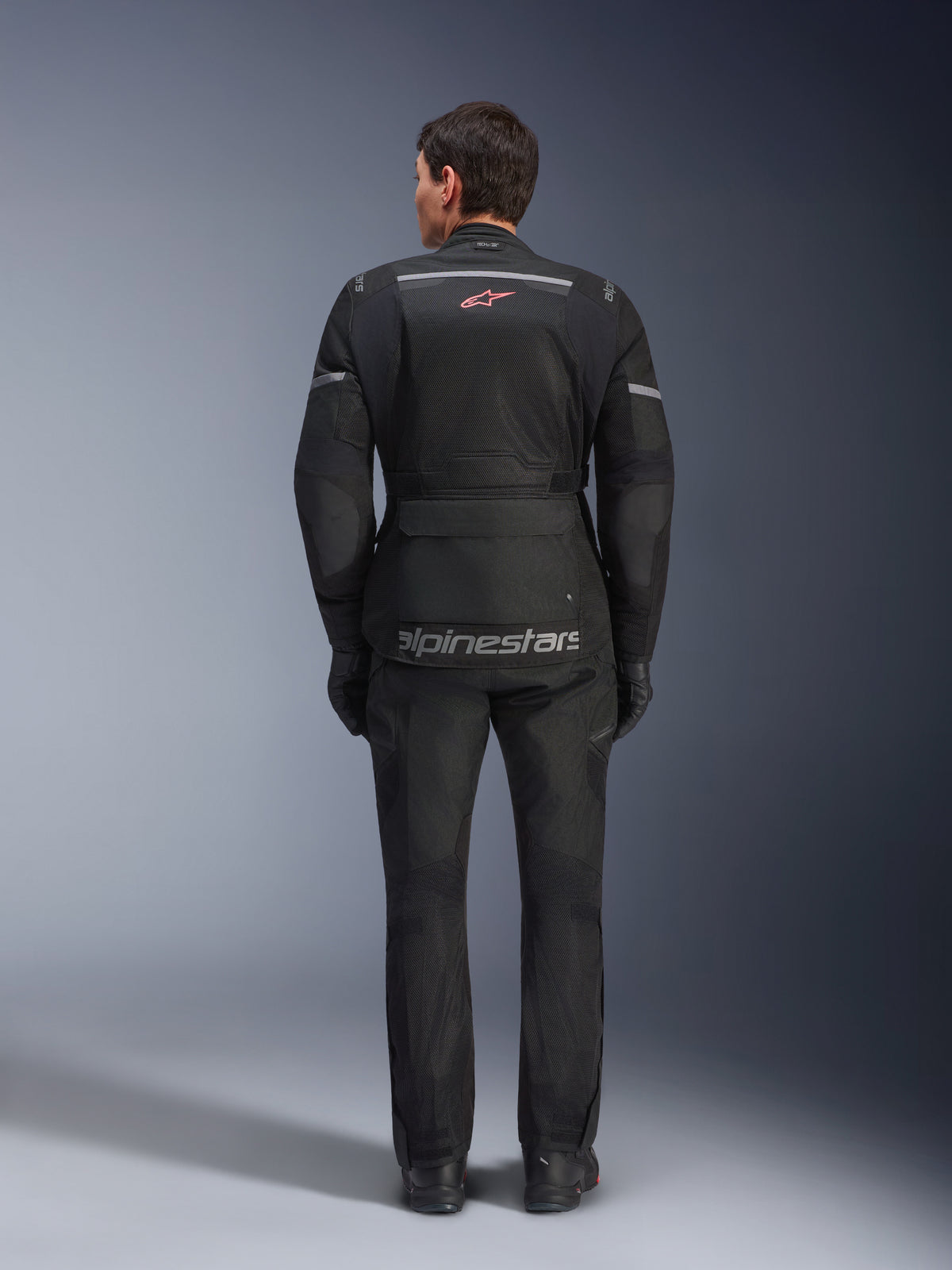 Kurtka i spodnie Alpinestars Andes Air Drystar®, czarny motocyklowy strój turystyczny, widok od tyłu na motocyklistę, panele z siatki, czerwone logo gwiazdy i odblaskowy napis Alpinestars na kurtce, w zestawie z czarnymi rękawicami i butami