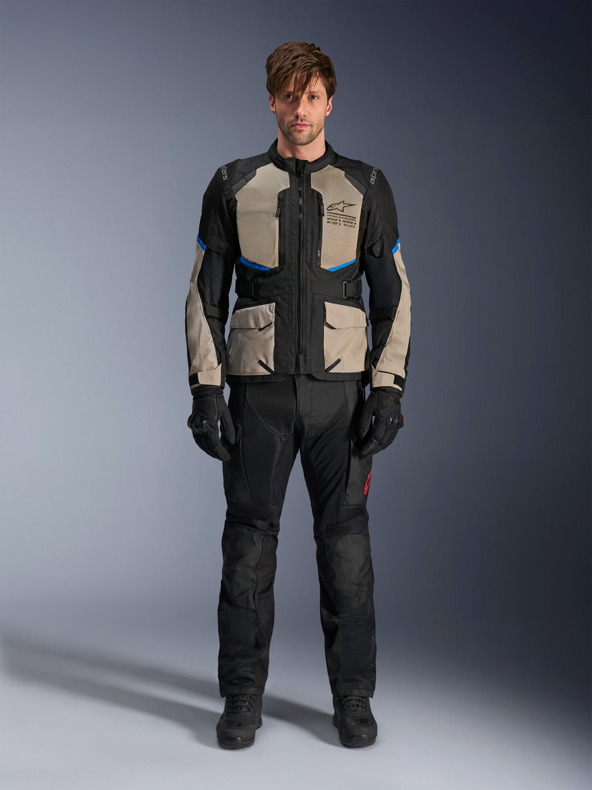 Model ubrany w kurtkę Alpinestars Andes Air Drystar®, motocyklowa kurtka turystyczna, kolor Black Laurel Gray i Bright Blue, zestawiona z pasującymi czarnymi spodniami motocyklowymi i rękawicami, z rozległymi panelami wentylacyjnymi z siatki na klatce piersiowej i rękawach do turystyki przygodowej w ciepłe dni.